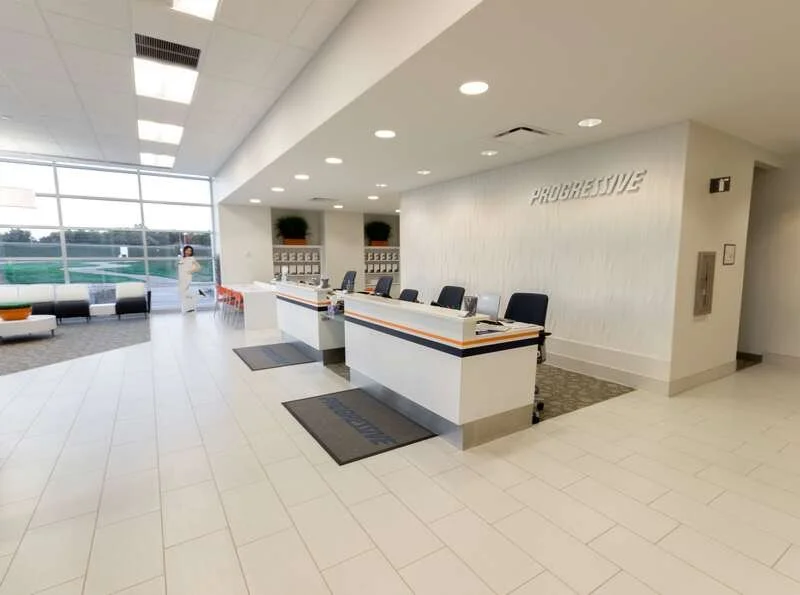 Modern office reception area with three check-in counters, black chairs, a person in white near a large window, and a wall with the word "PROGRESSIVE" on it.
