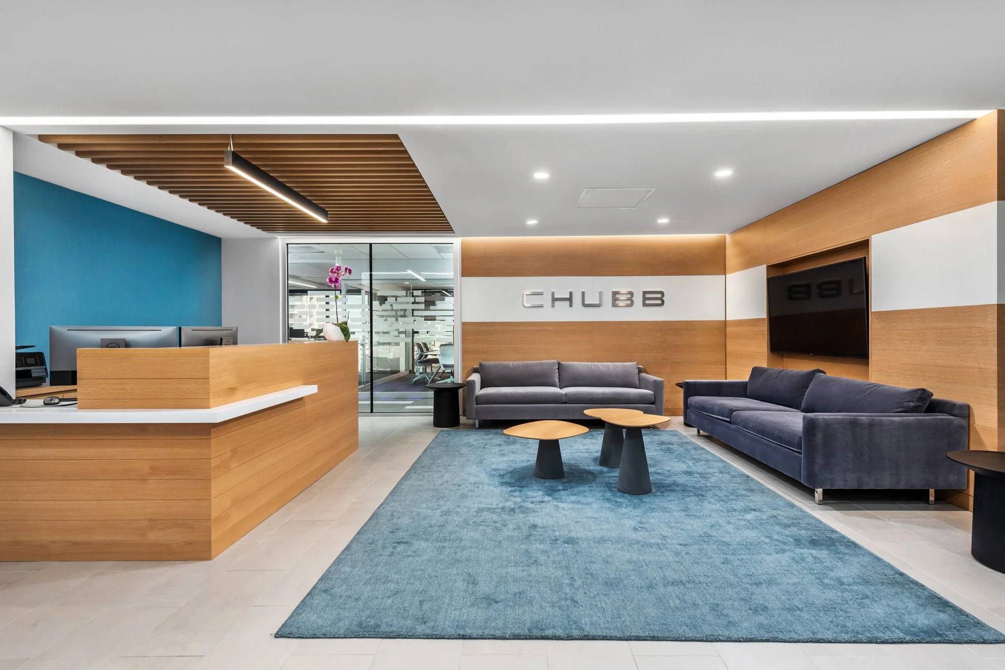 Modern office lobby featuring a reception desk to the left, two sofas in gray and navy blue facing each other with two wooden coffee tables in between, a blue rug, a TV mounted on a wooden wall to the right, and a glass door leading to an adjacent conference room in the background. The decor includes a purple orchid on the reception desk and the company name 'CHUBB' on the wall behind the sofas.