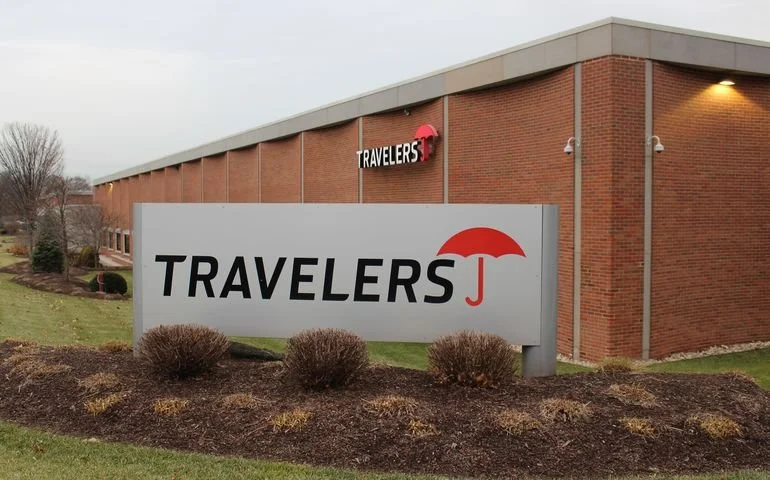 Travelers Insurance building with a large sign featuring the company name and a red umbrella logo, surrounded by bushes and grass.