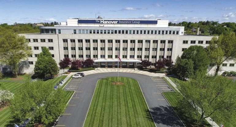 A large office building with a sign that reads 'The Hanover Insurance Group' on top. The building has multiple floors with many windows and is surrounded by well-maintained trees and a parking lot. There is a grassy area and a small circular driveway in front with an American flag.