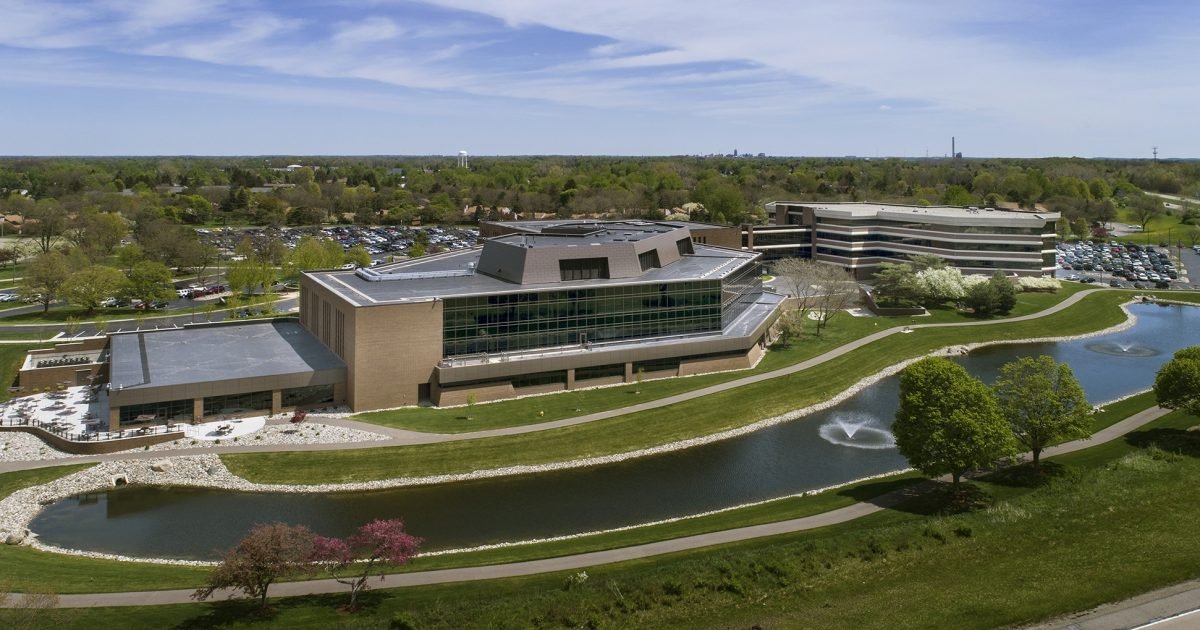 Aerial view of a modern office building complex surrounded by a landscaped park with a pond, trees, and walking paths.