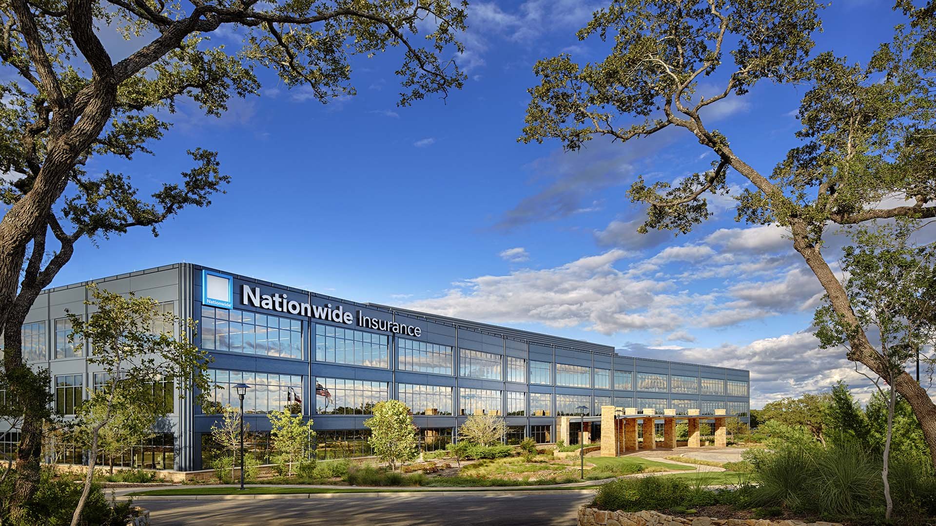 Exterior view of a modern, multi-story office building with a sign that reads 'Nationwide Insurance'; the building has large glass windows reflecting a blue sky with scattered clouds, surrounded by trees and landscaped greenery.