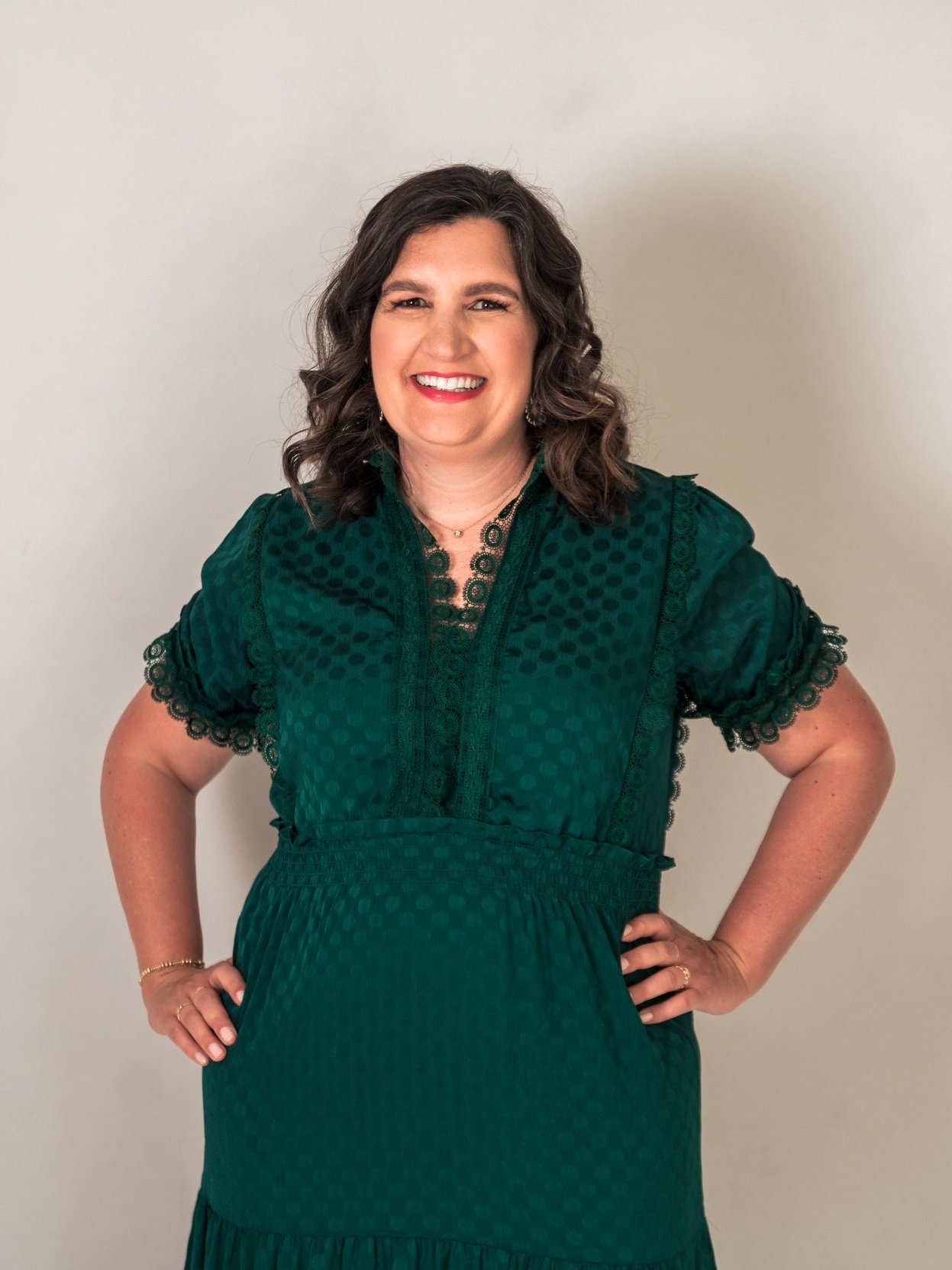 Smiling woman in a green dress with lace details, hands on hips, against a neutral background.