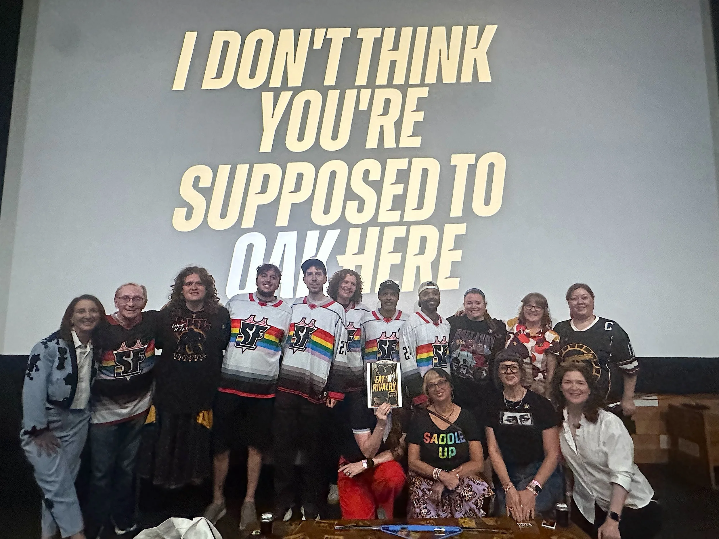 Color photo of 15 people in costumes, HR gear, and Quakes hockey merch, standing and kneeling in front of a movie theater screen bearing ghte event logo for "I DON'T THINK YOU'RE SUPPOSED TO OAK HERE."