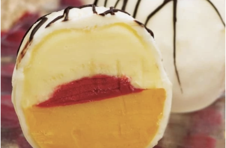 Close-up of a sliced dessert with layers of yellow, red, and white, topped with chocolate drizzle.