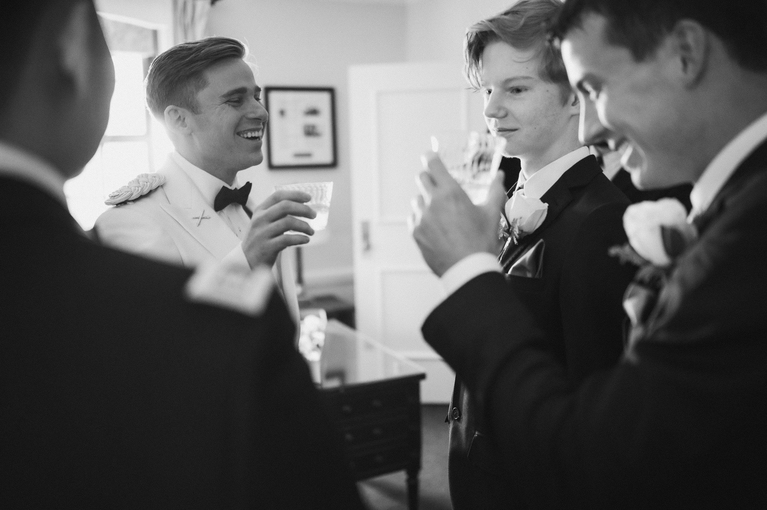 Four men in formal tuxedos and boutonnieres, holding glasses, smiling and chatting indoors at a wedding celebration.