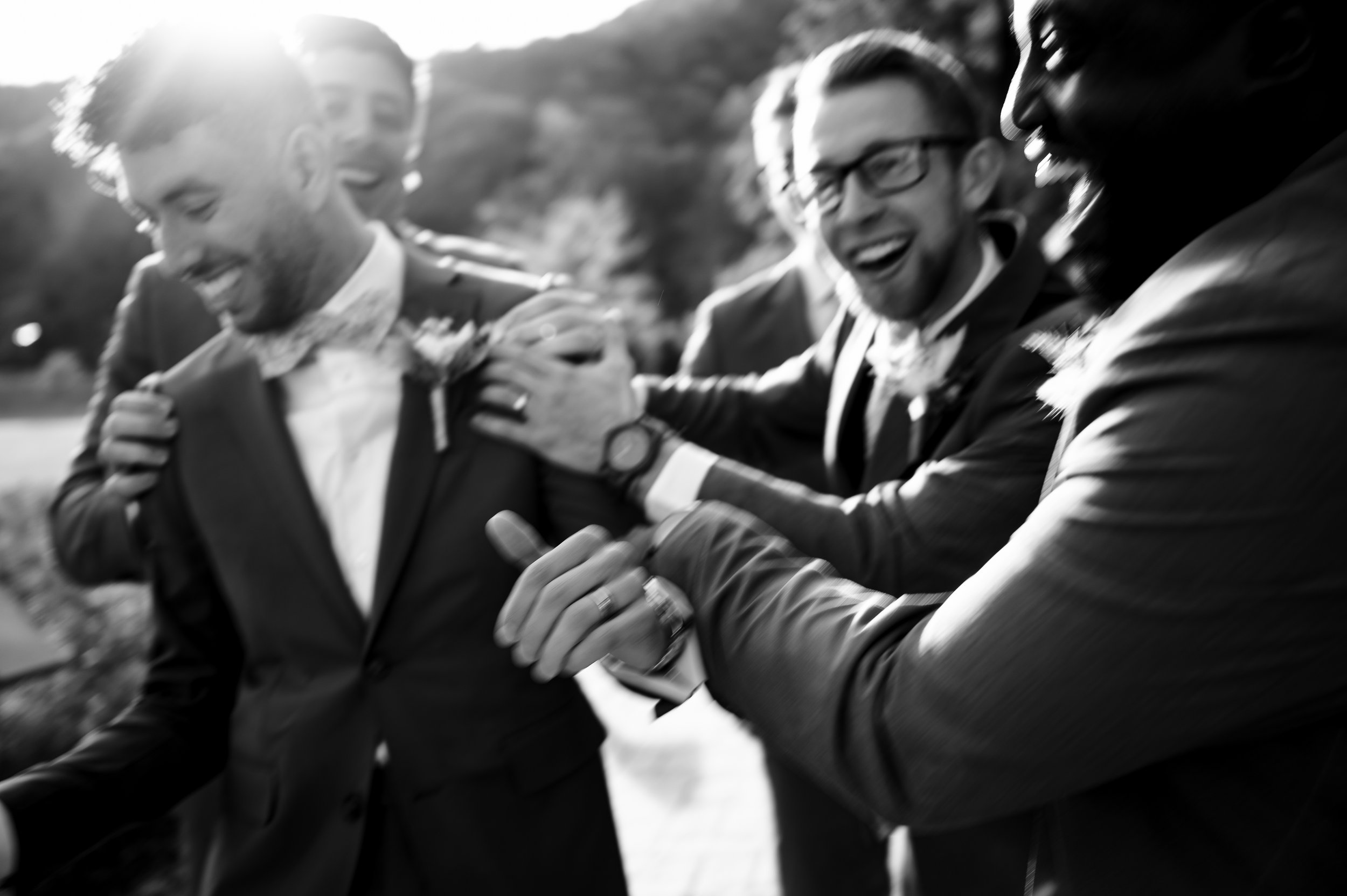 Group of men in suits and tuxedos, smiling and laughing together outdoors in sunlight, celebrating wedding or special occasion.