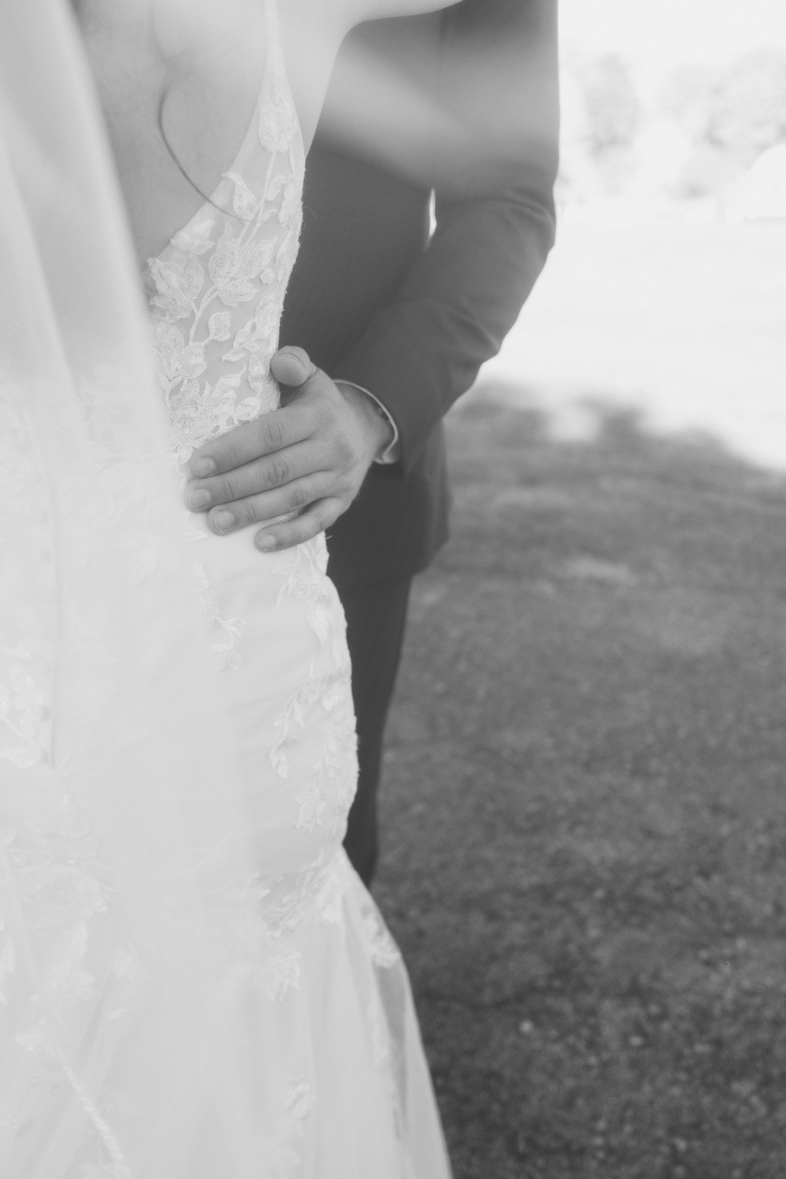 Close-up of a bride and groom holding hands, with the bride's hand on her waist, wearing wedding attire, outdoors.