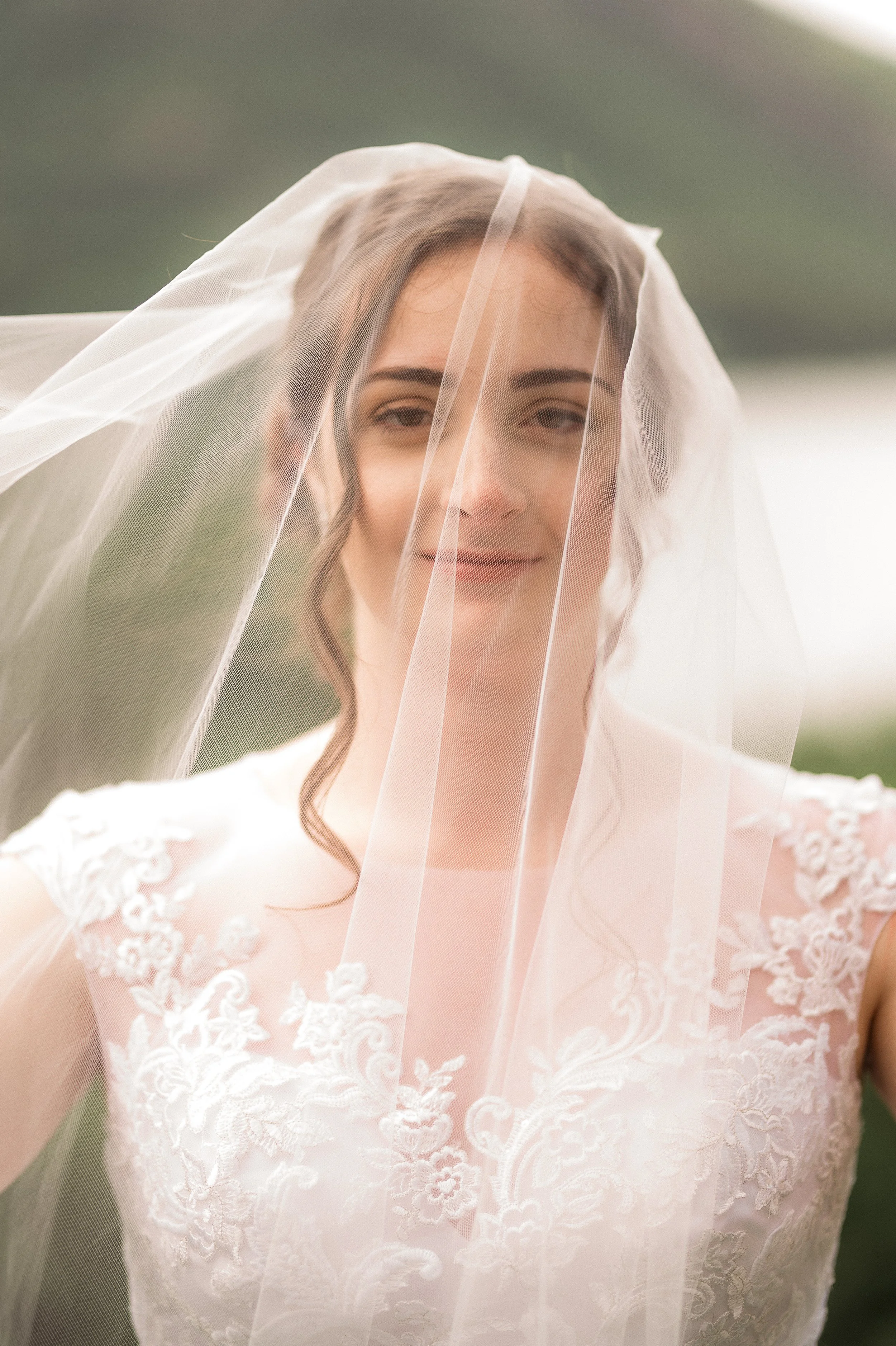 A bride with a veil on her head, smiling gently, wearing a lace wedding dress with floral embroidery.