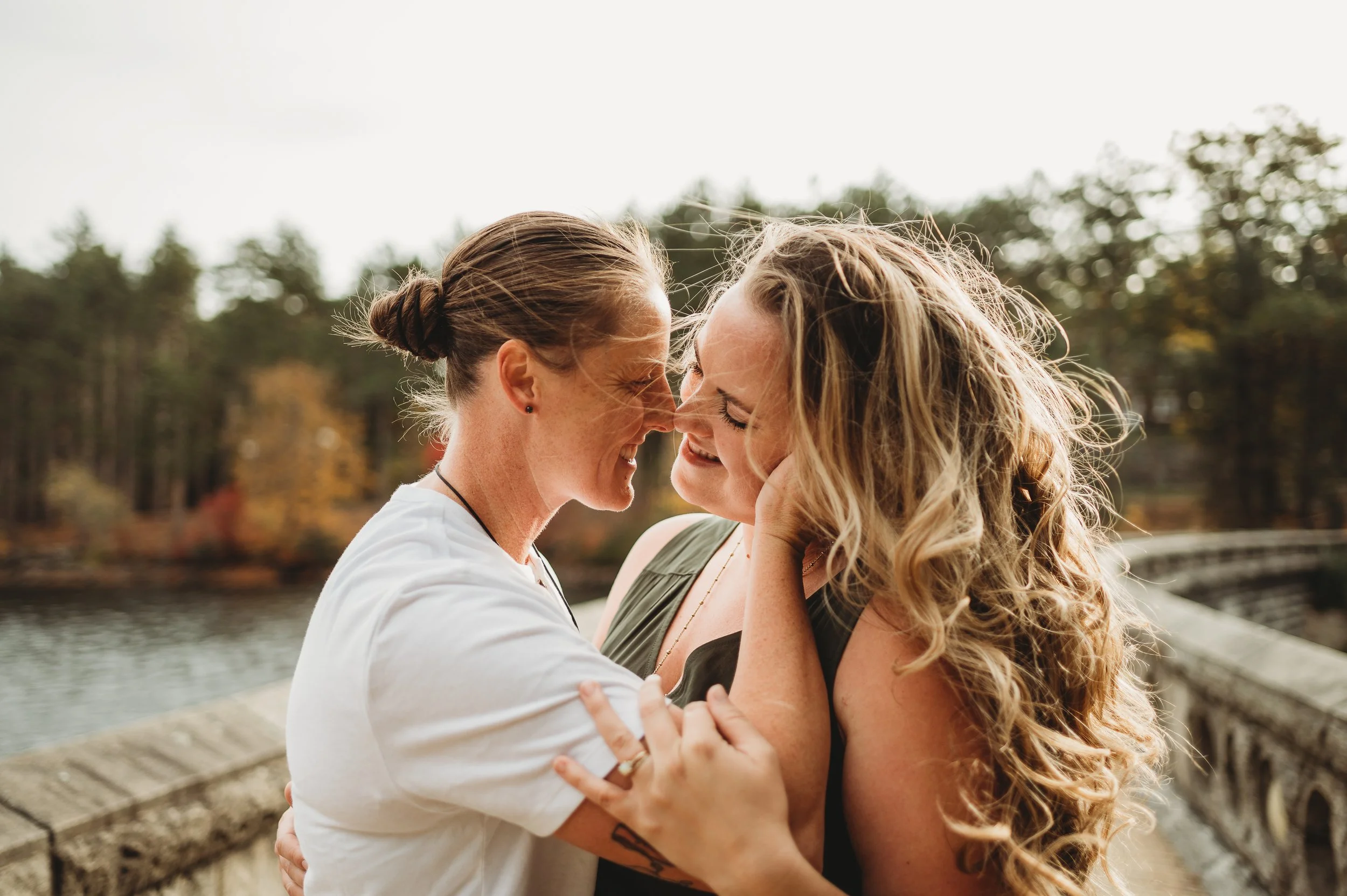 Two women are smiling and touching noses, embracing each other outdoors near a body of water with trees in the background.