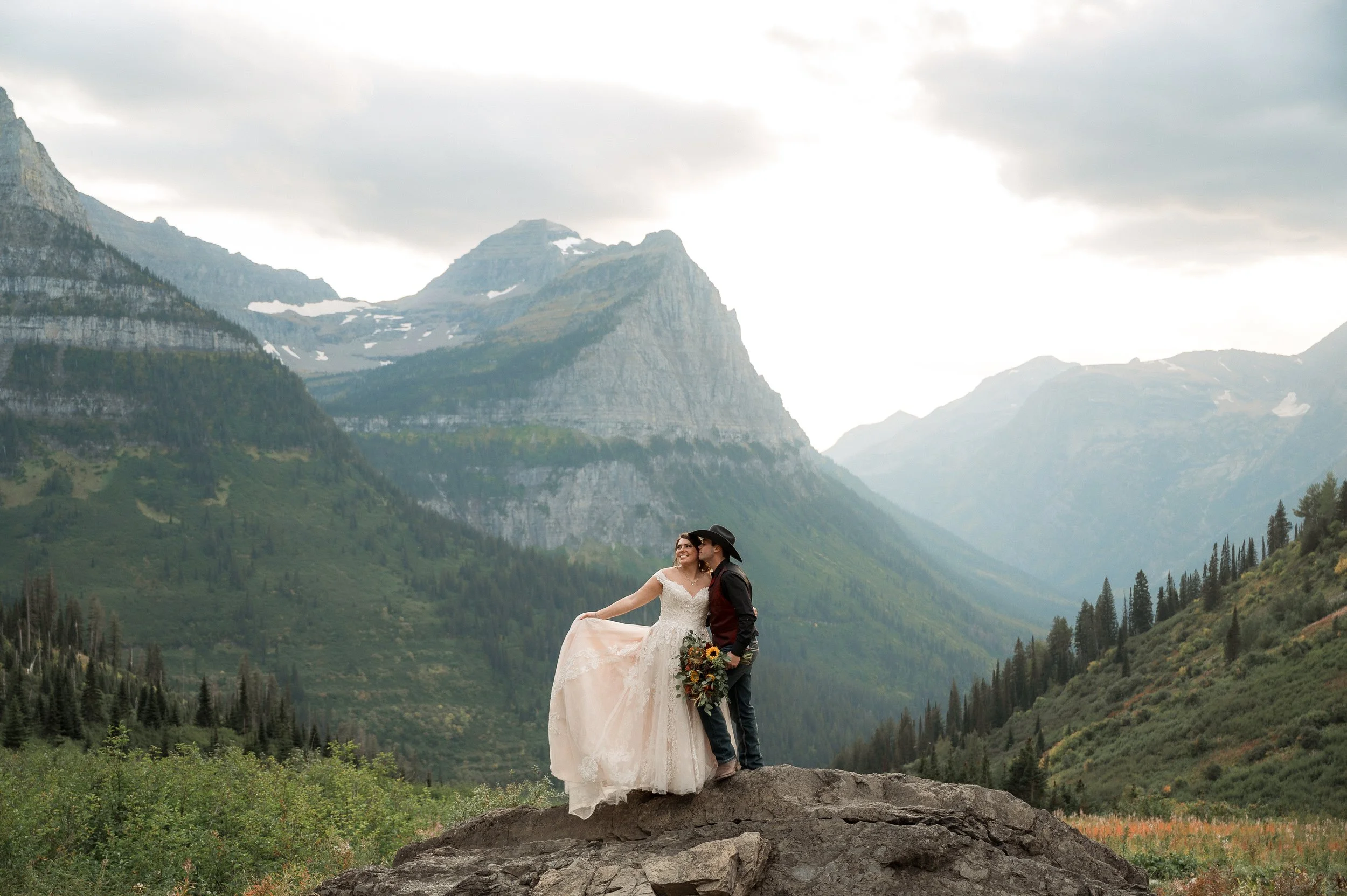 A couple wedding photo on a large rock in a mountainous landscape with green trees and snow-capped peaks in the background.