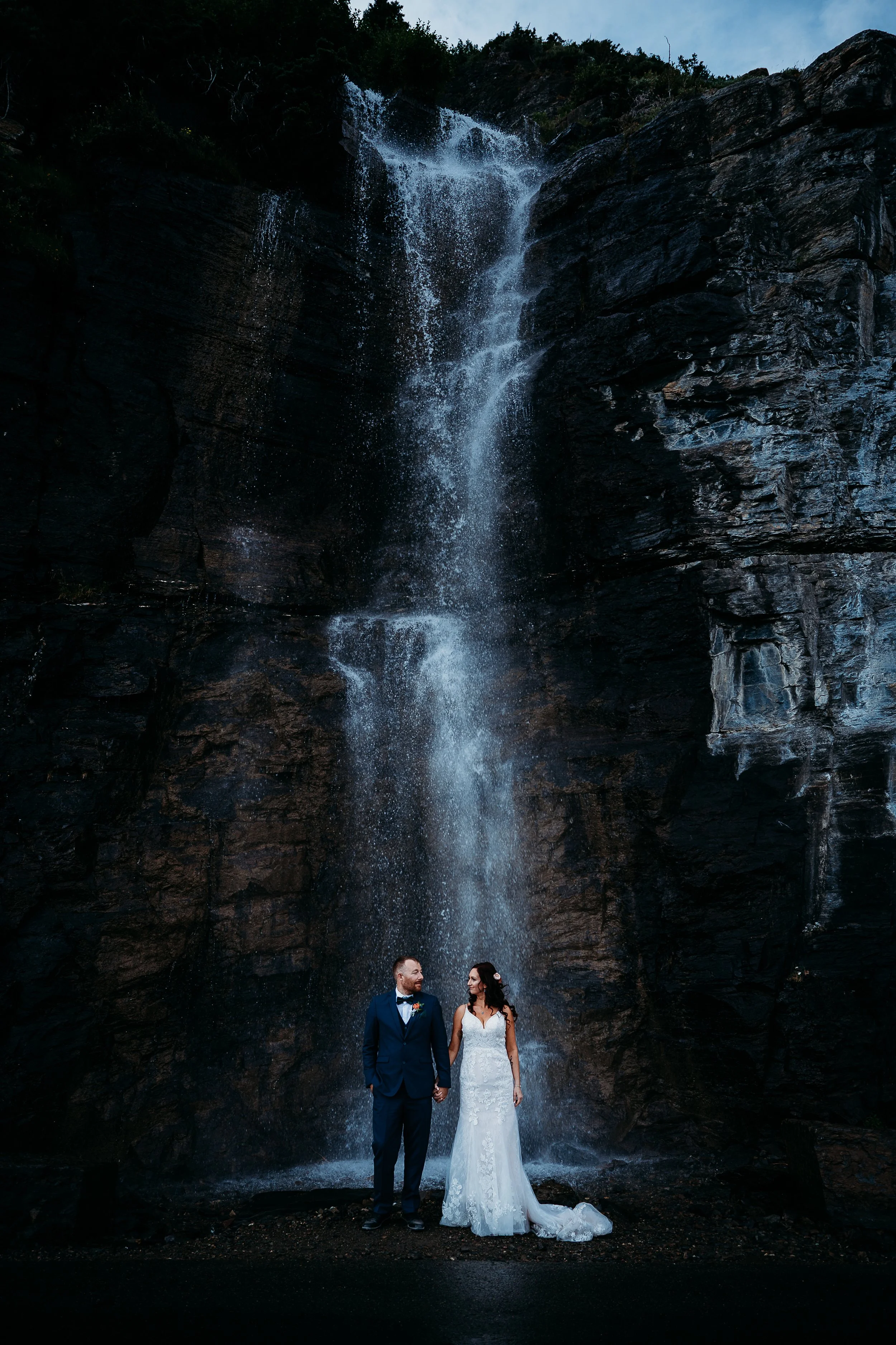 A bride and groom holding hands and smiling at each other in front of a tall waterfall, with dark rocks and greenery around.