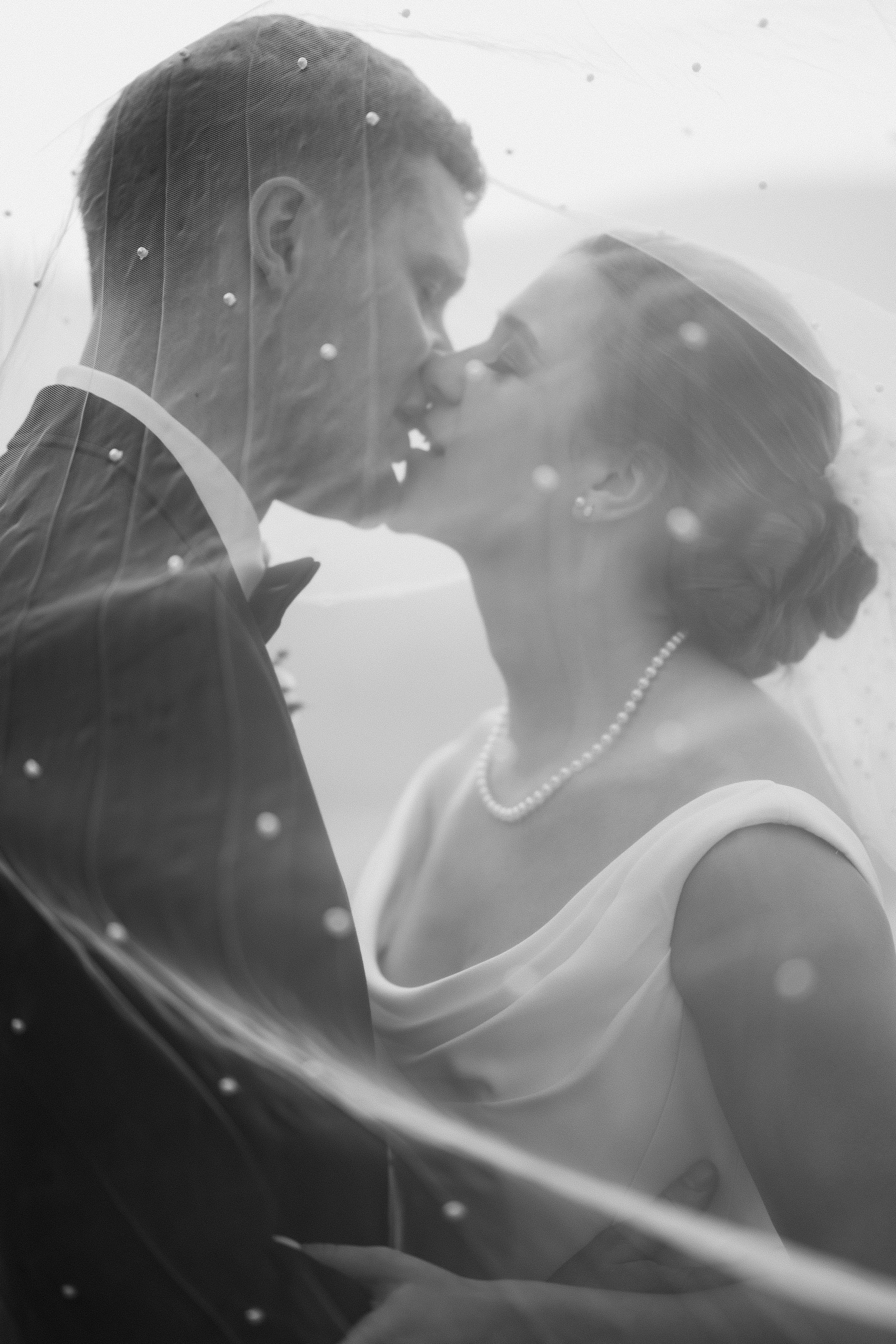 Black and white photograph of a bride and groom sharing a kiss, taken through a wedding veil decorated with small pearls.