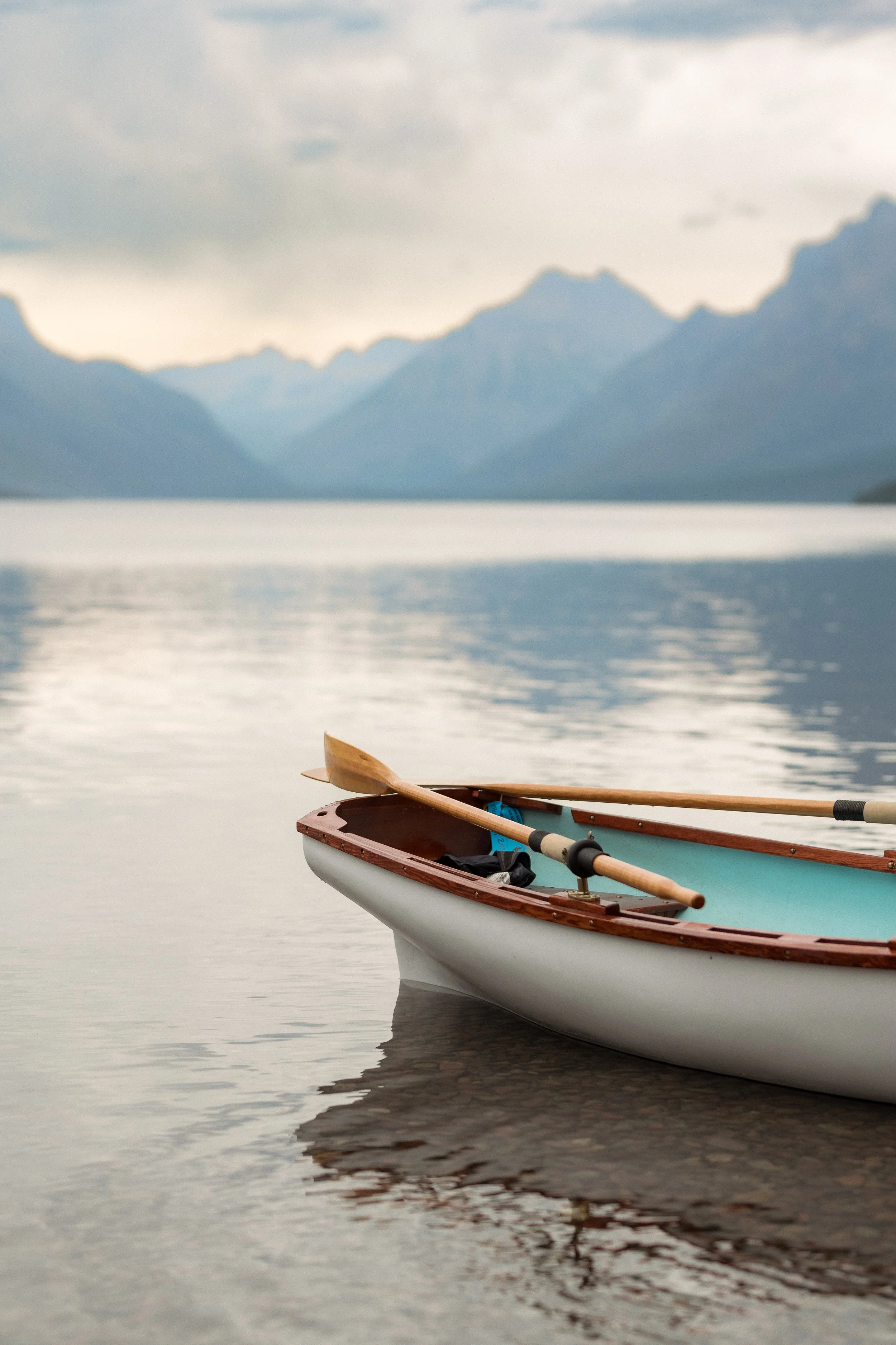 A rowboat with oars floats on a calm lake with mountains and cloudy sky in the background.