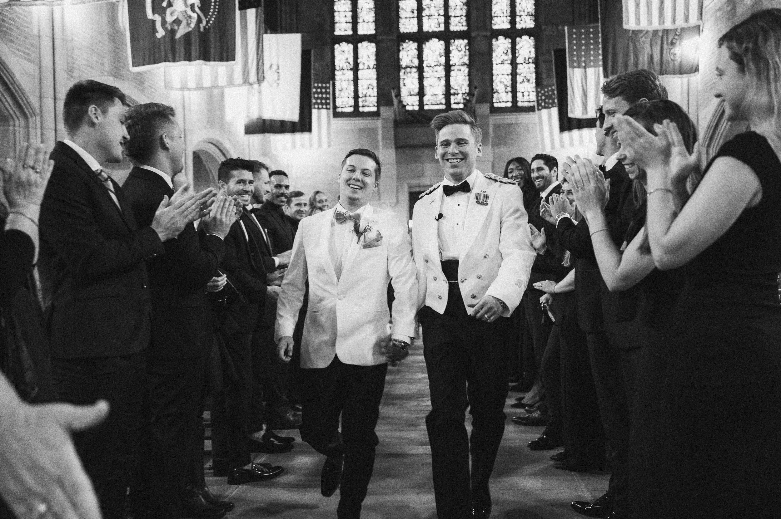Two men in white tuxedos, one with medals, walking hand in hand down an aisle in a church, surrounded by smiling guests clapping and cheering.