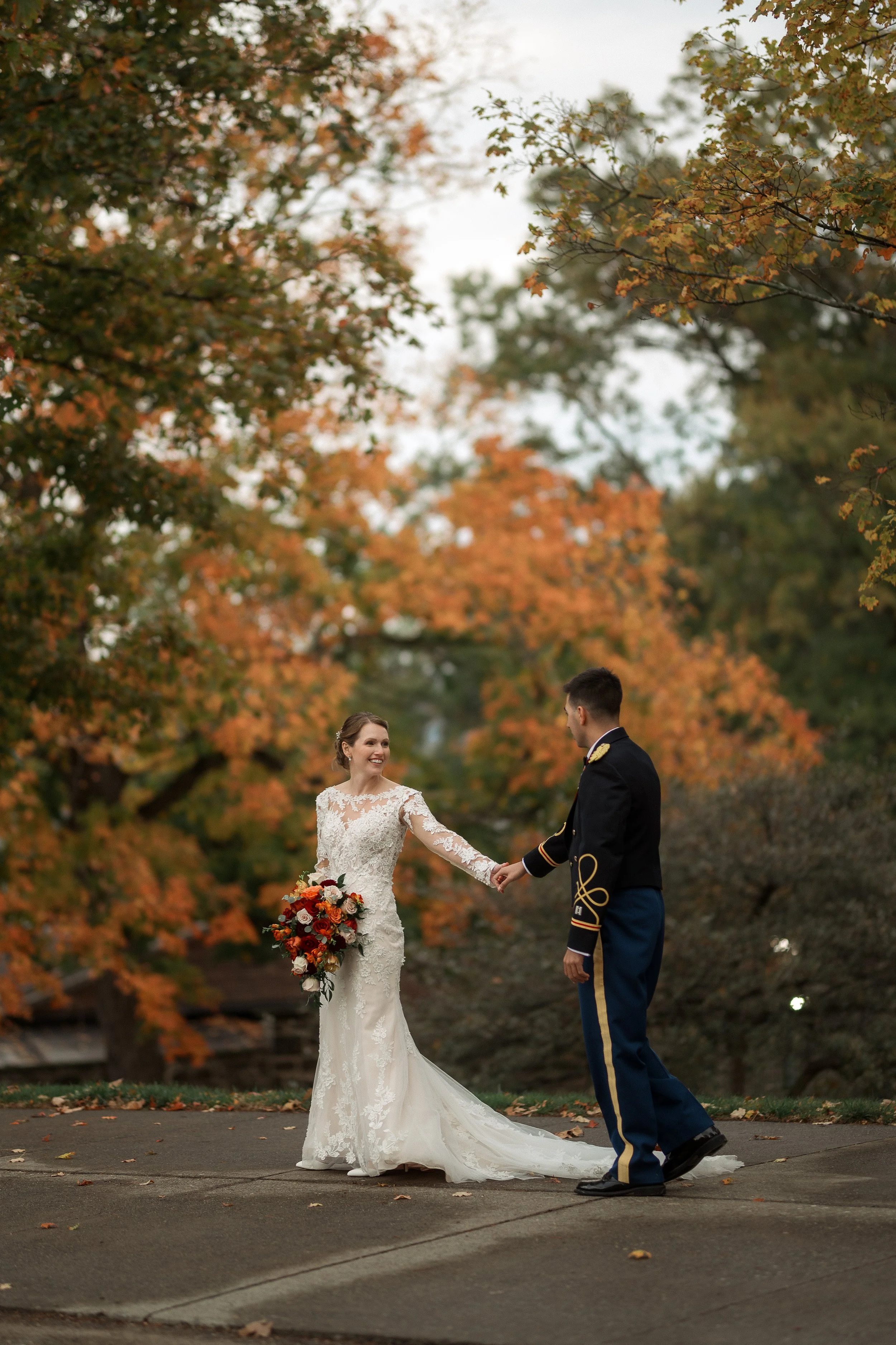 A bride and groom holding hands outdoors during fall, with trees in orange and green leaves in the background.