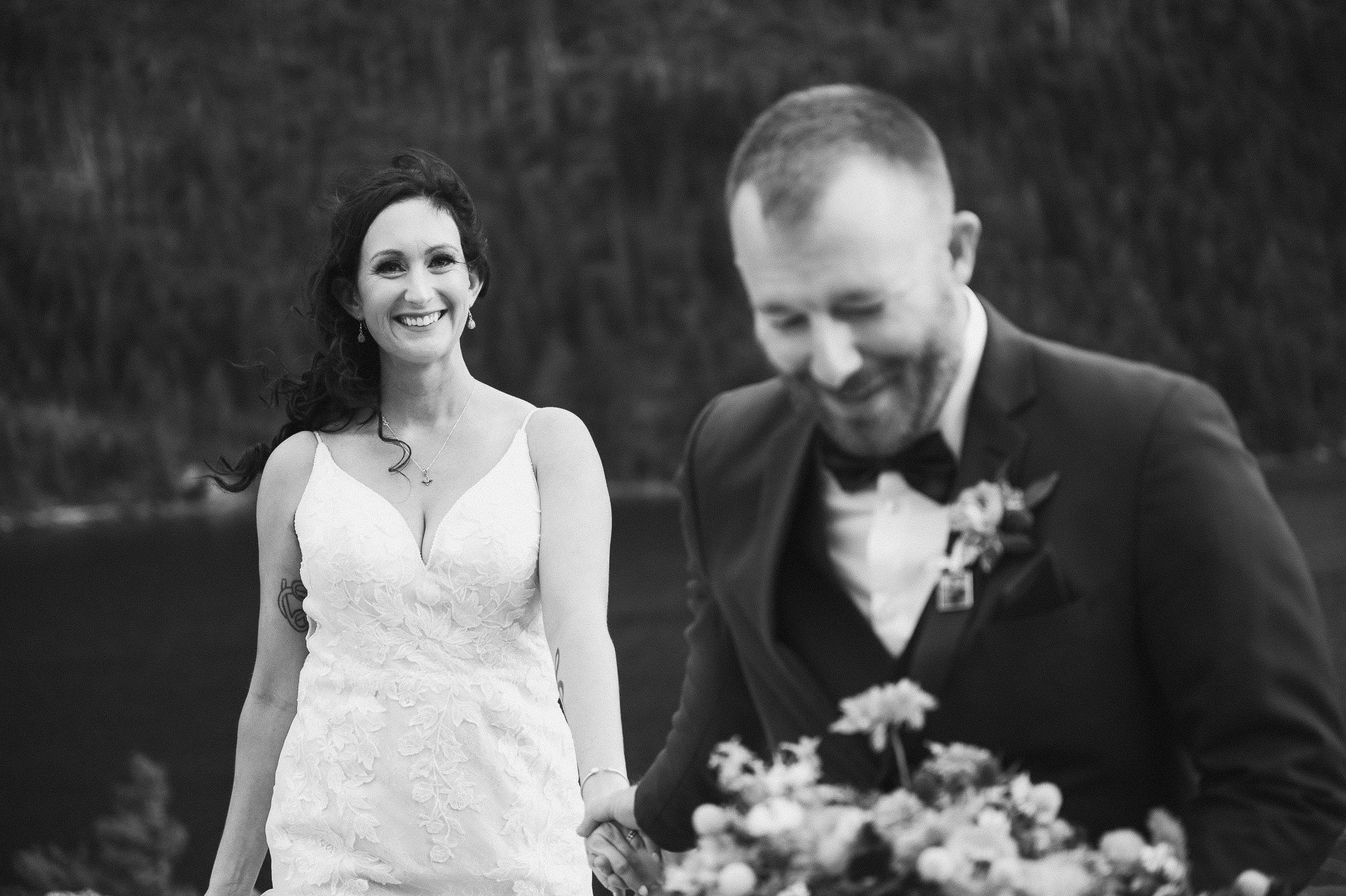 A smiling woman in a white wedding dress stands near a man in a tuxedo holding a bouquet. They are outdoors near a body of water and trees.