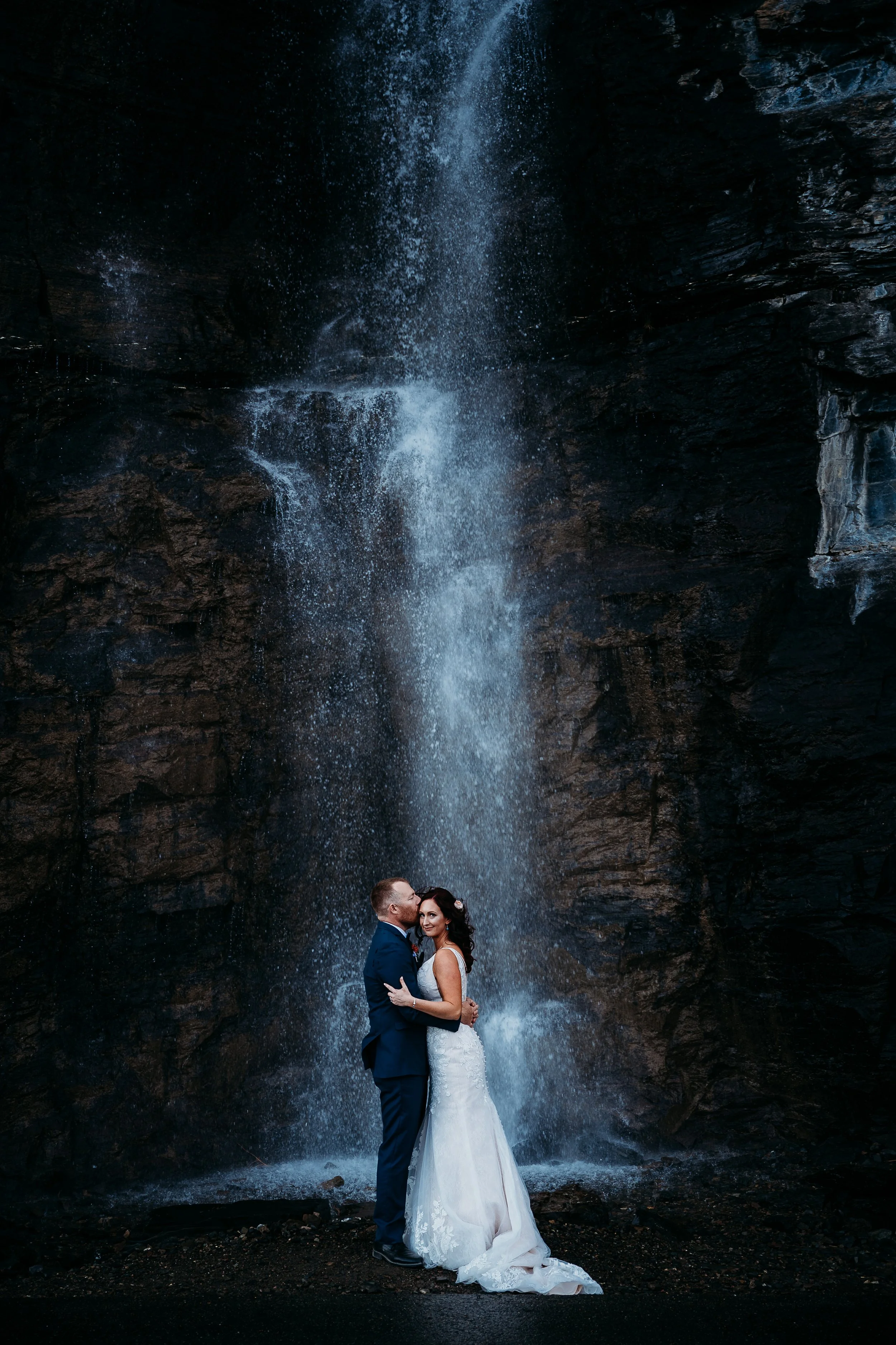 A bride and groom in wedding attire embracing in front of a tall waterfall.