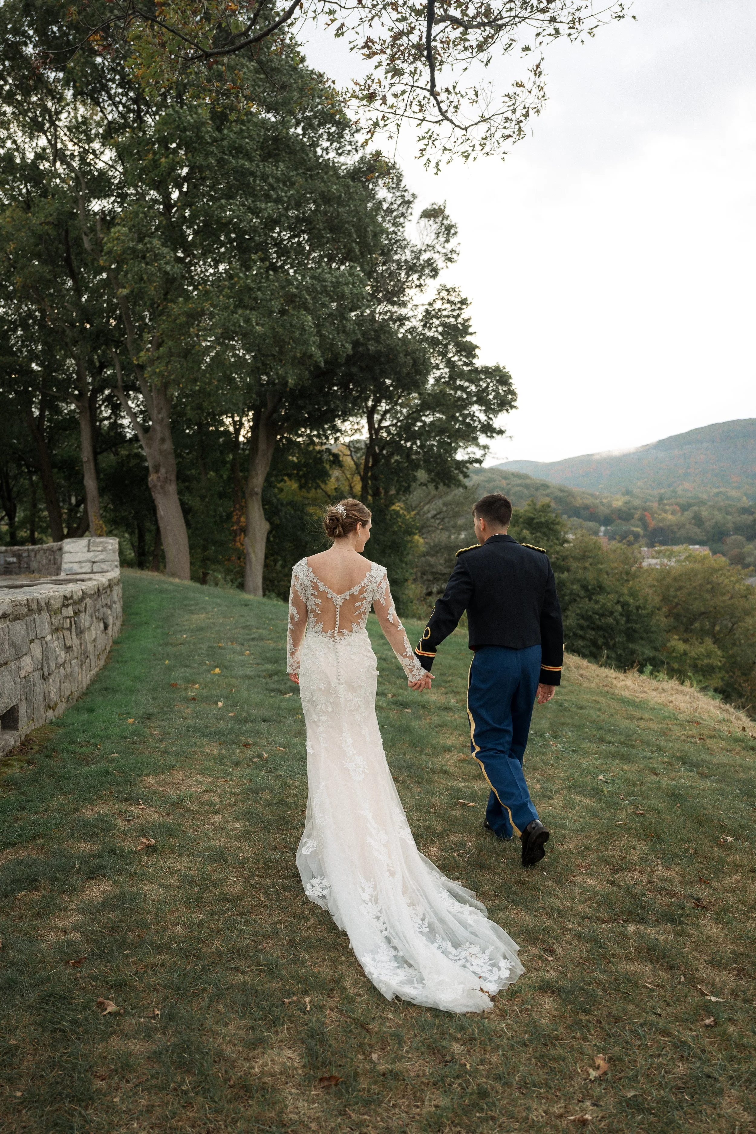 A bride and groom walking hand in hand outdoors on a grassy hillside with trees and mountains in the background, holding hands with their backs to the camera.