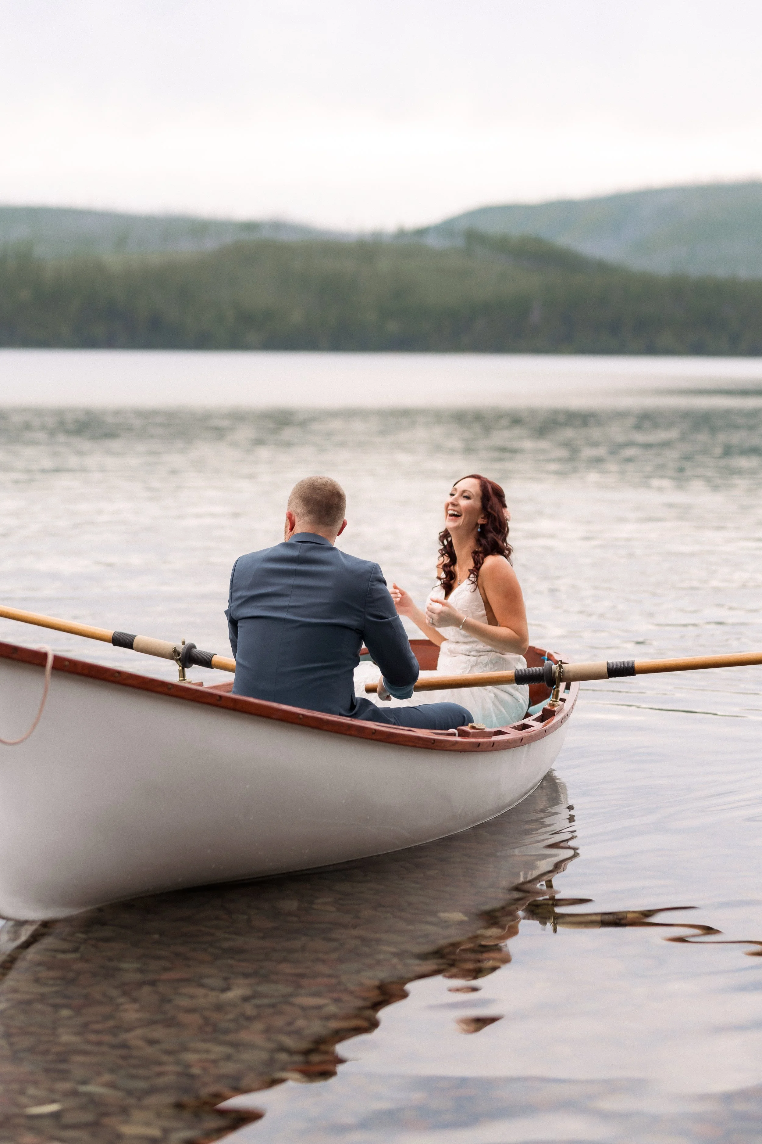 A couple in wedding attire sitting in a rowboat on a calm lake, surrounded by mountain scenery, with the bride laughing.