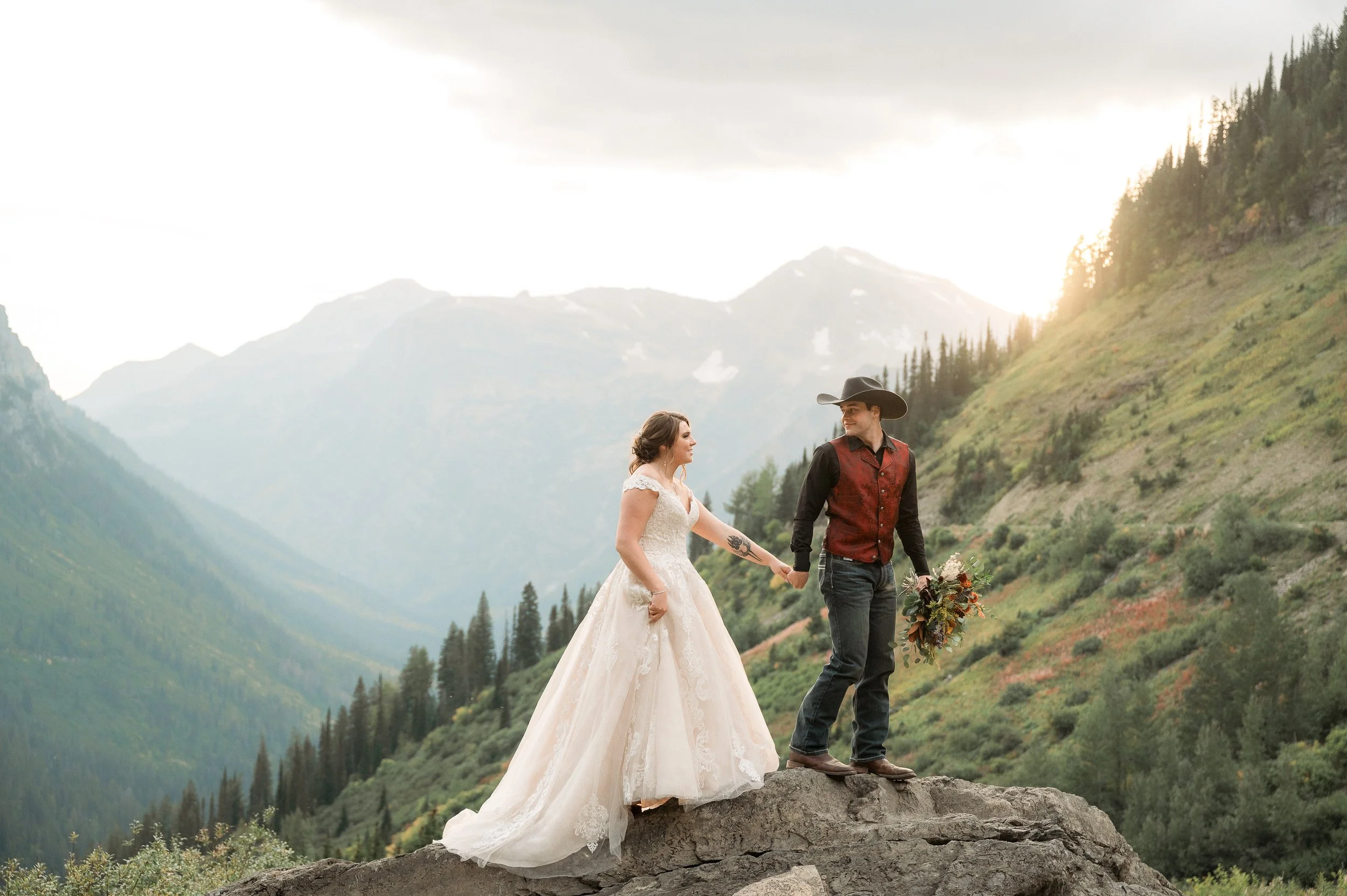 A newlywed couple holds hands on a rocky ledge outdoors in a mountainous landscape at sunset. The bride wears a white lace wedding gown, and the groom wears a cowboy hat, a western-style vest, and jeans, holding a bouquet of flowers.