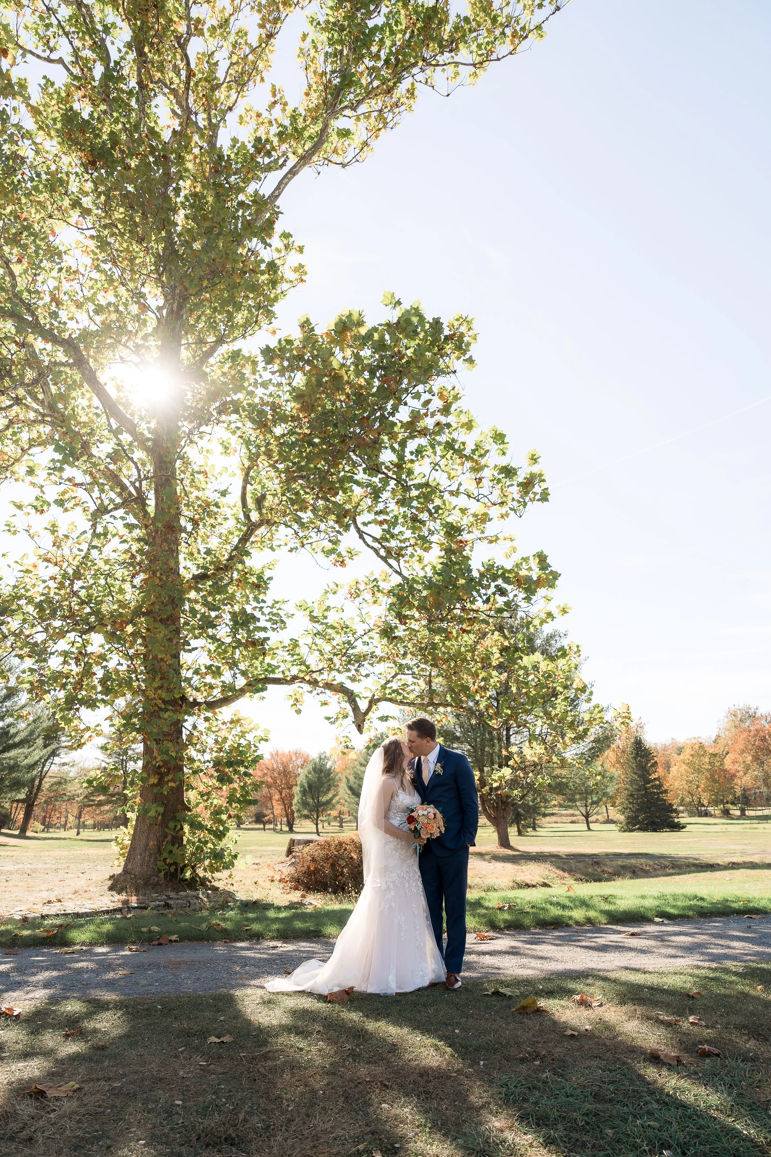 A bride and groom share a kiss outdoors under a large tree on a sunny day, surrounded by autumn foliage.