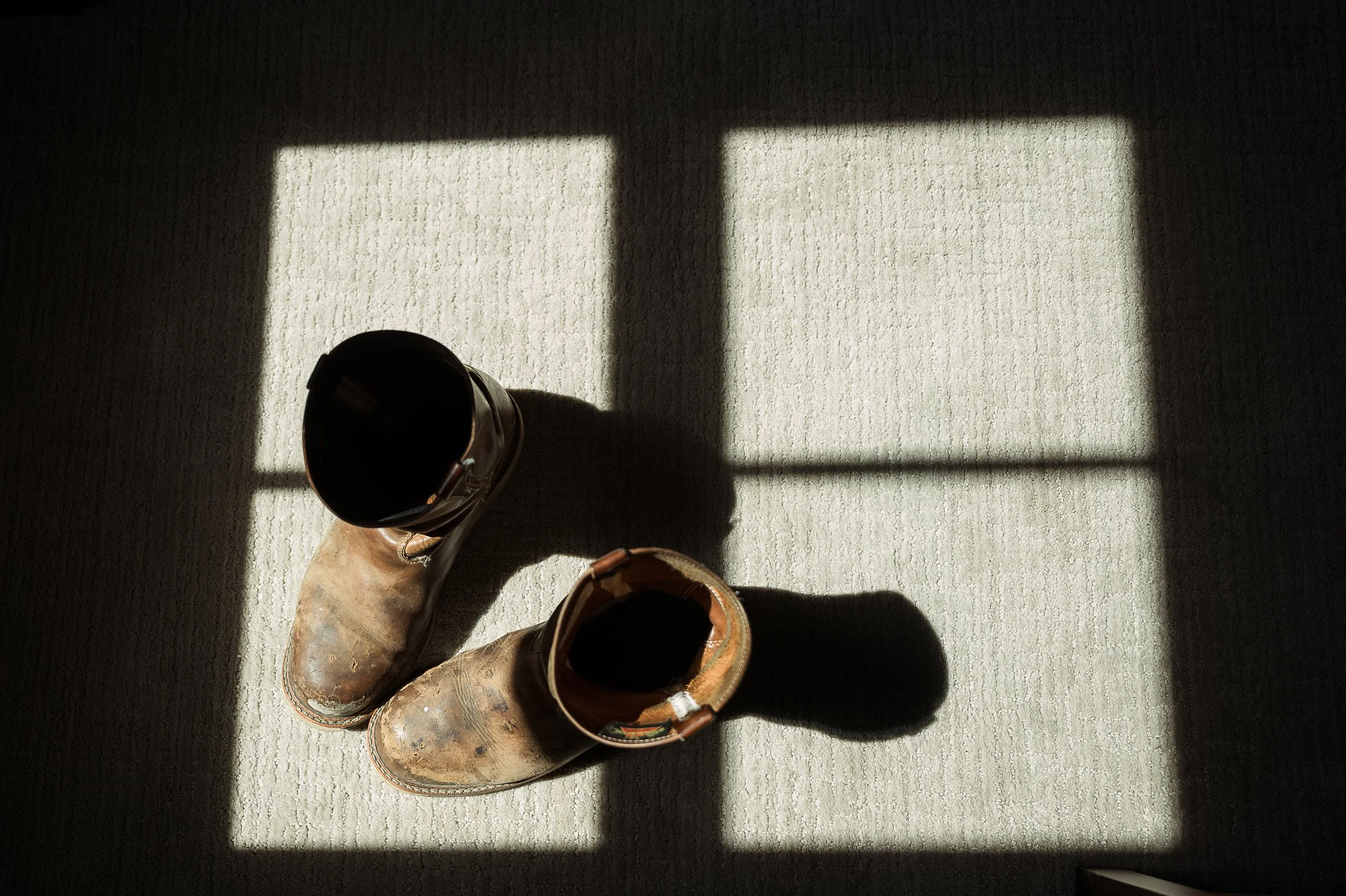 Pair of worn cowboy boots on beige carpet with sunlight coming through a window, creating a shadow.