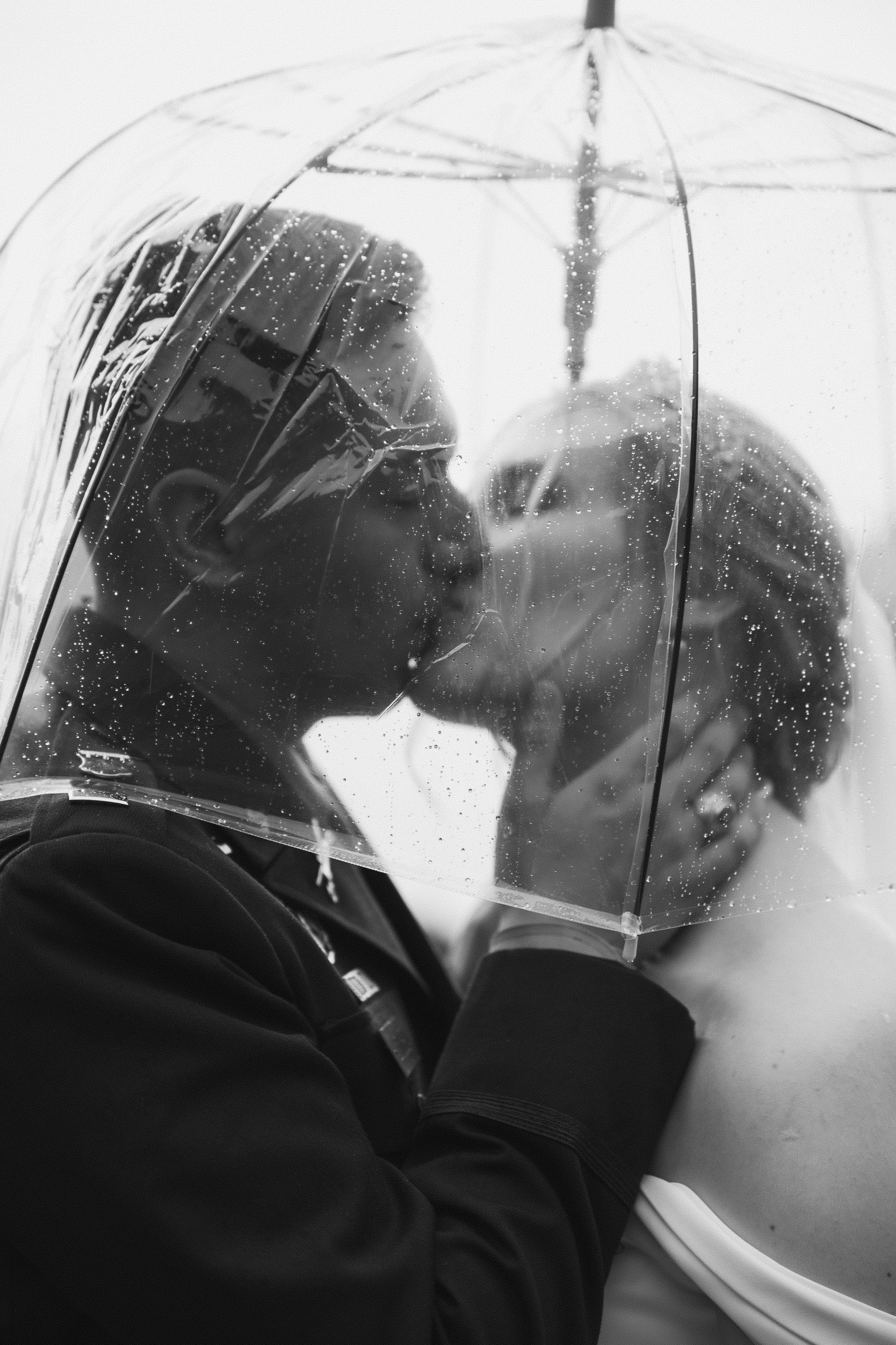 A black-and-white photo of a couple kissing under a transparent umbrella on a rainy day.