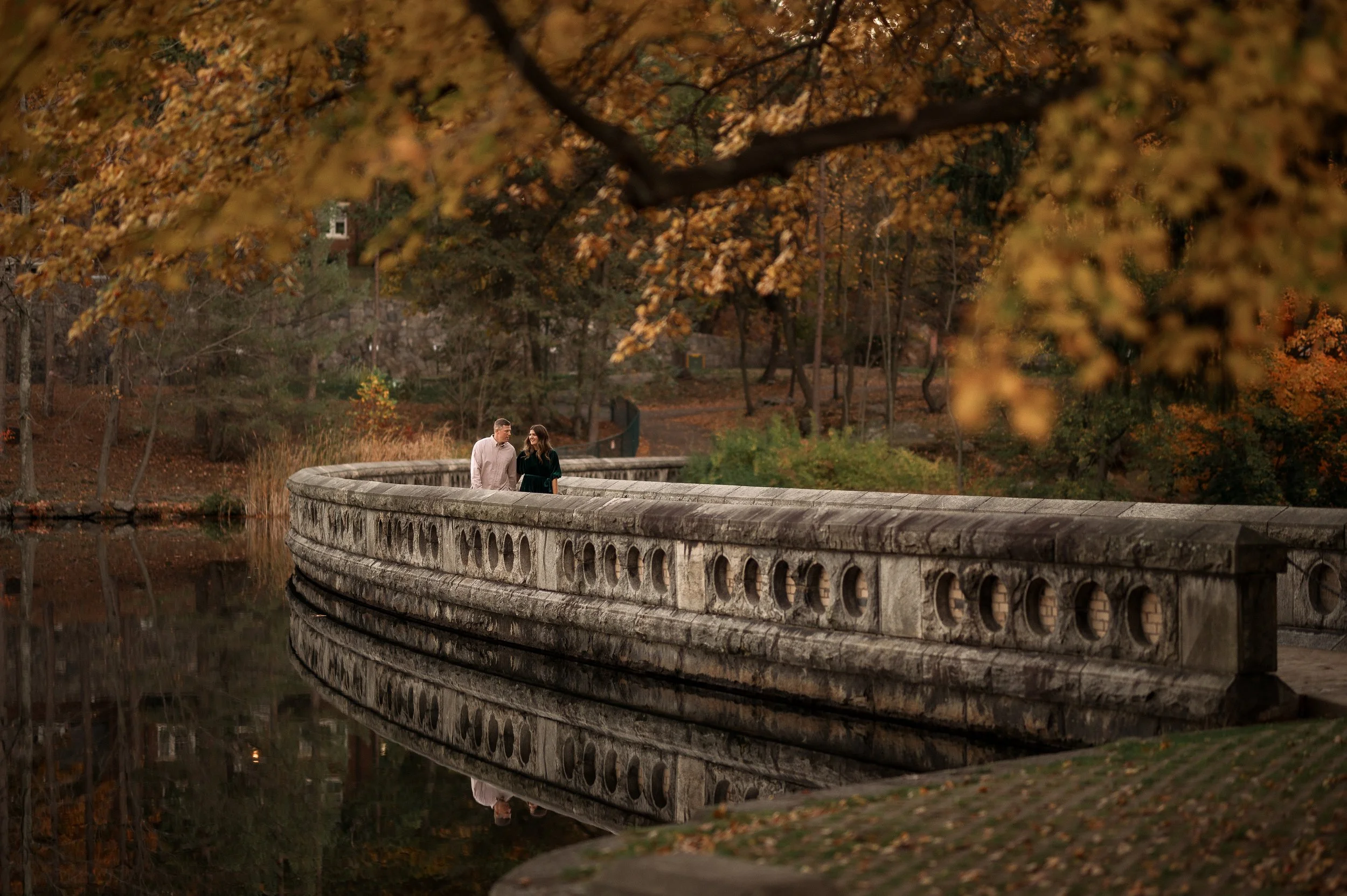 A man and woman walking on a curved stone bridge over a calm body of water, with autumn trees and leaves in the background.