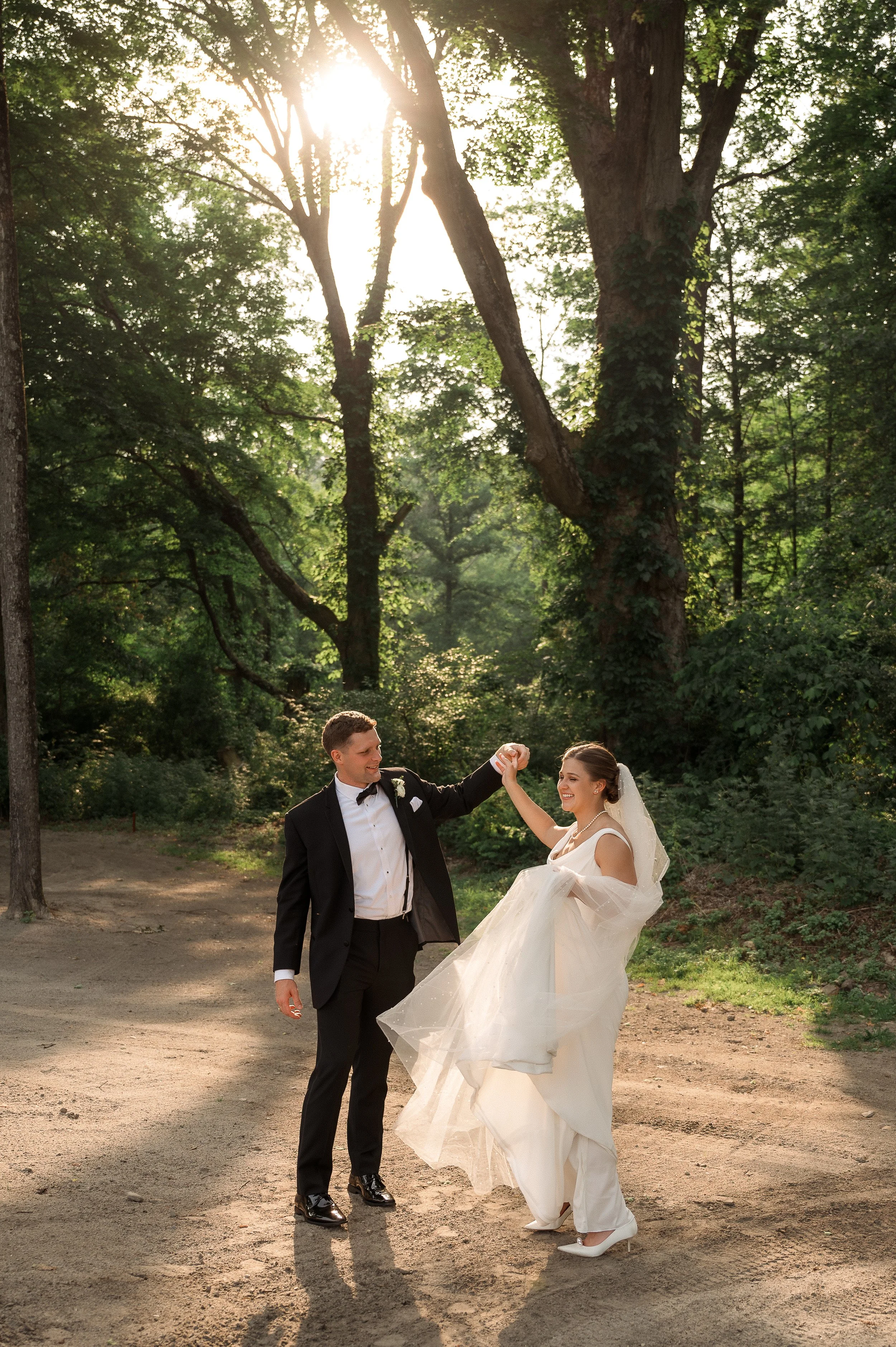 A bride and groom dancing outdoors on a dirt path in a wooded area during sunset, with sunlight filtering through trees.