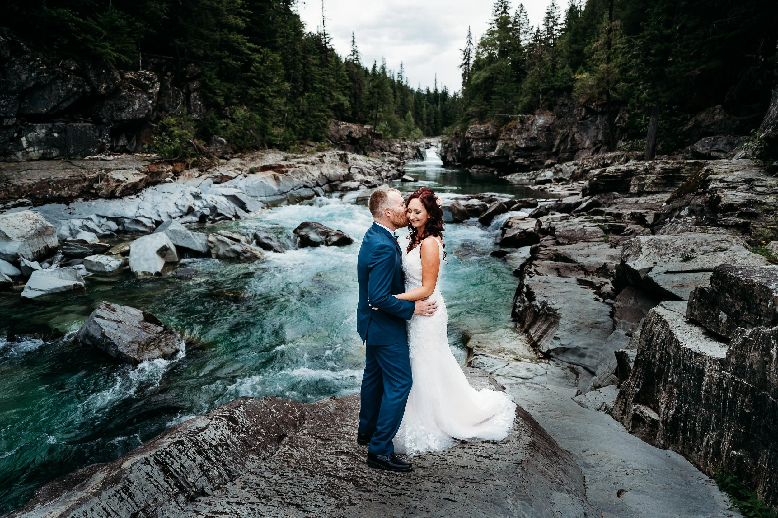 A newlywed couple in wedding attire sharing a kiss on a large rock in a mountain river canyon surrounded by evergreen trees.