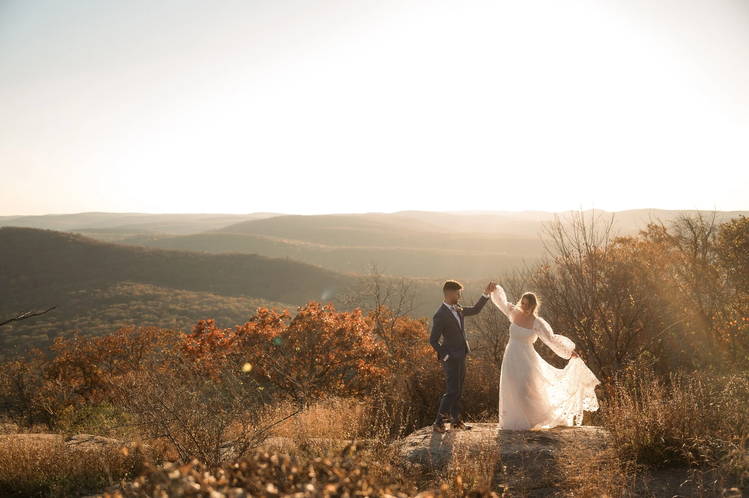 A bride and groom dancing on a large rock in a scenic outdoor setting with rolling hills and autumn trees during sunset.