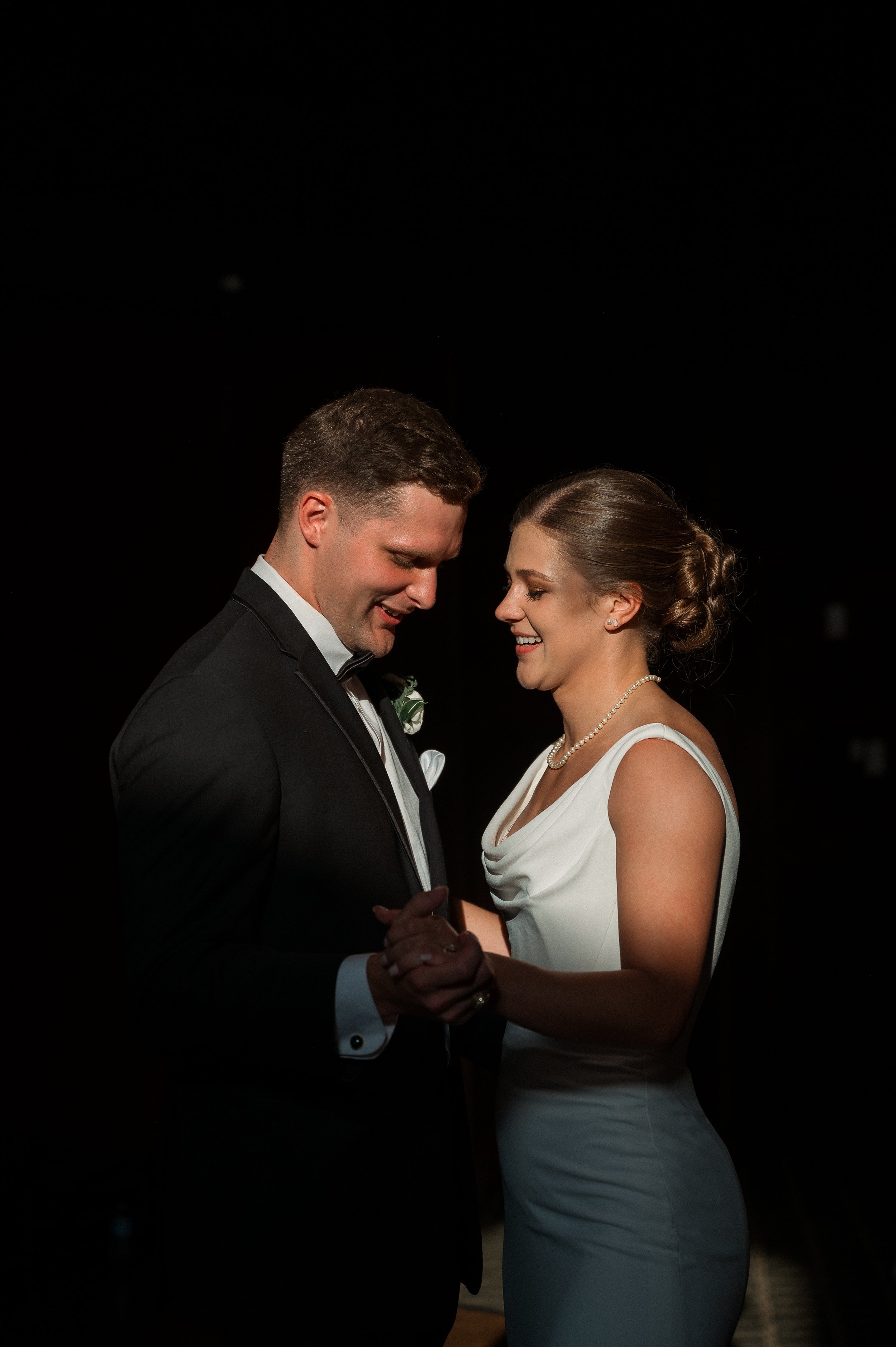 A newlywed couple dancing at their wedding reception, smiling and holding hands in a dark setting.