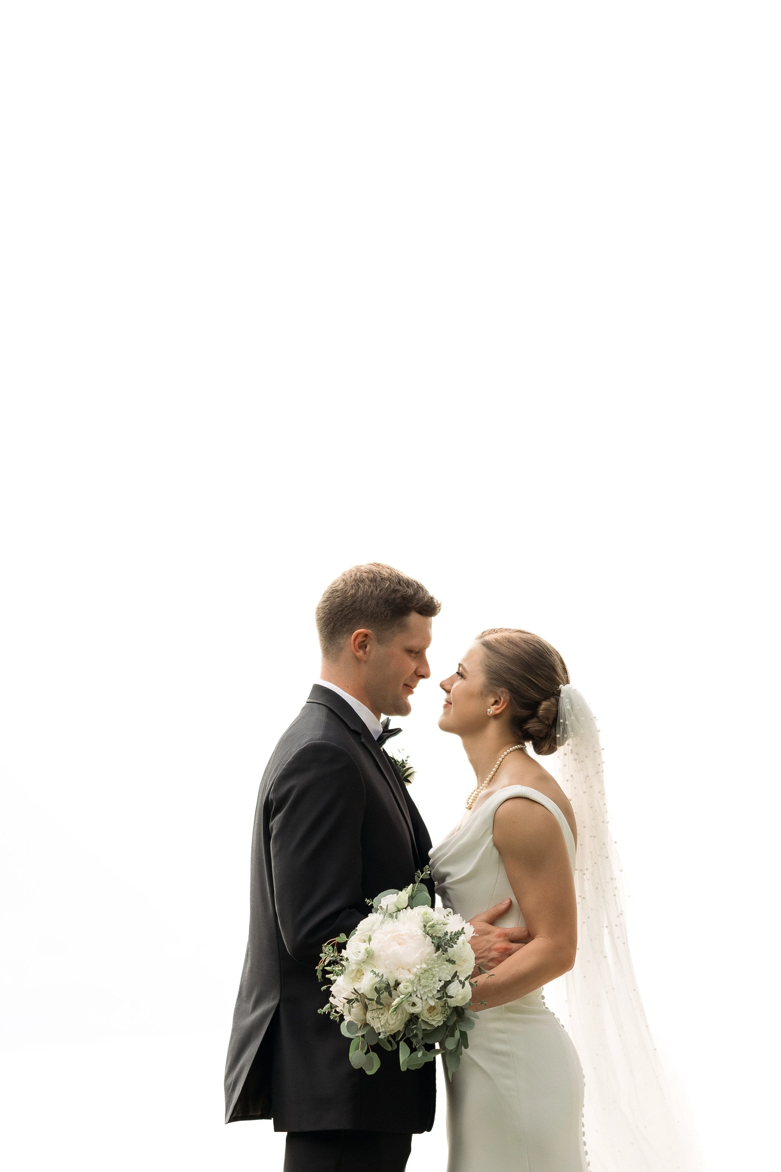 A bride and groom standing close together, gazing into each other's eyes on their wedding day. The bride holds a bouquet of white flowers, and the groom wears a black tuxedo with a bow tie.