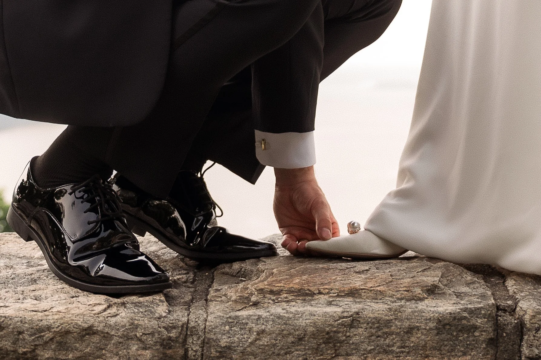 Close-up of a wedding ceremony where a person in a black suit and shiny black shoes is kneeling on a stone surface, adjusting a white shoe on another person's foot dressed in a white gown.