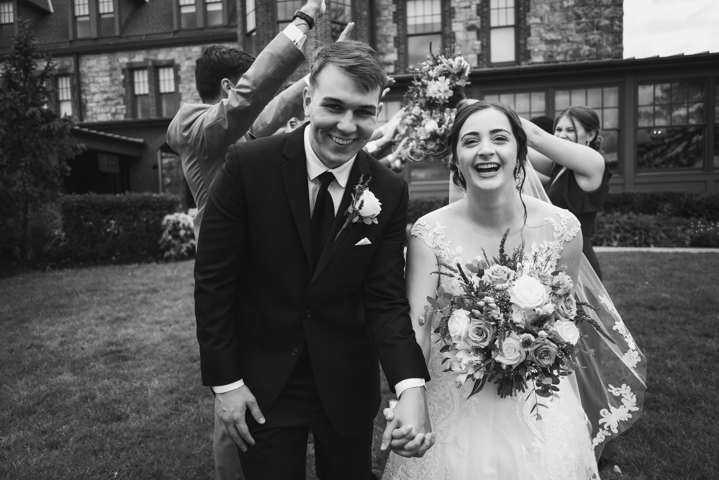 Black and white photo of a newly married couple holding hands and smiling, with wedding party members behind them throwing flower petals, in an outdoor garden area.