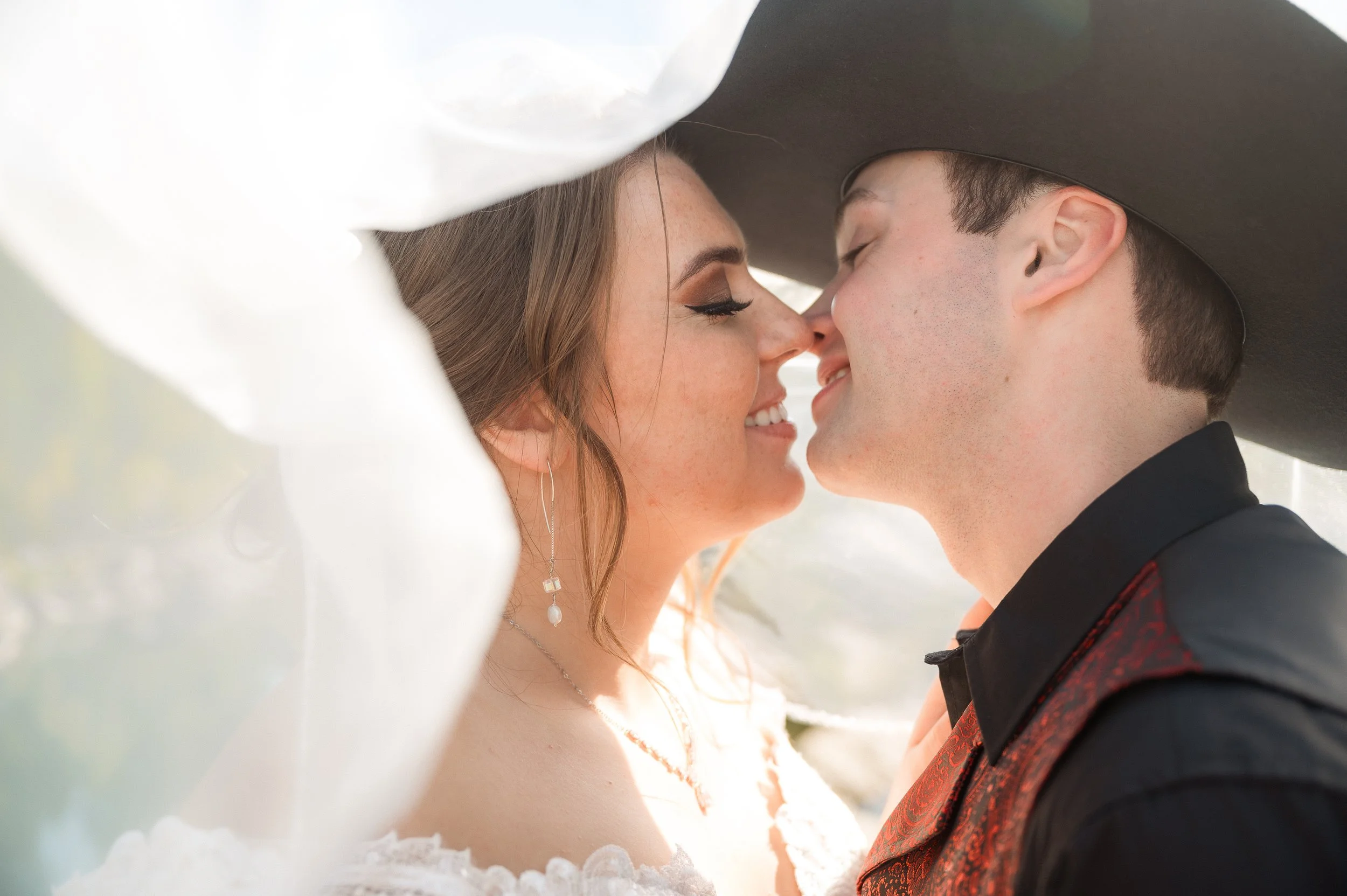 A couple in wedding attire close to each other with noses touching, under the bride's veil and wearing traditional Western wedding clothes.