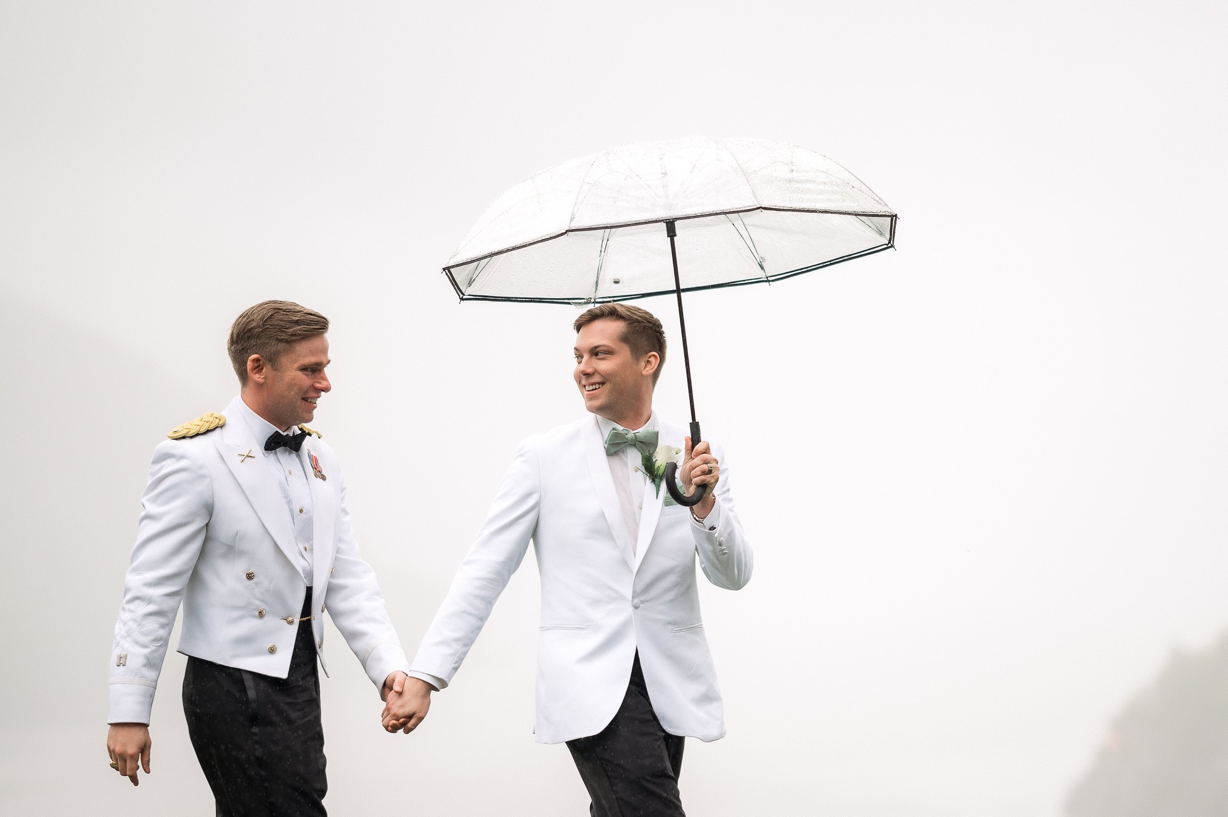 Two men dressed in wedding attire, one with a military uniform, holding hands under a clear umbrella on a cloudy day, smiling at each other.