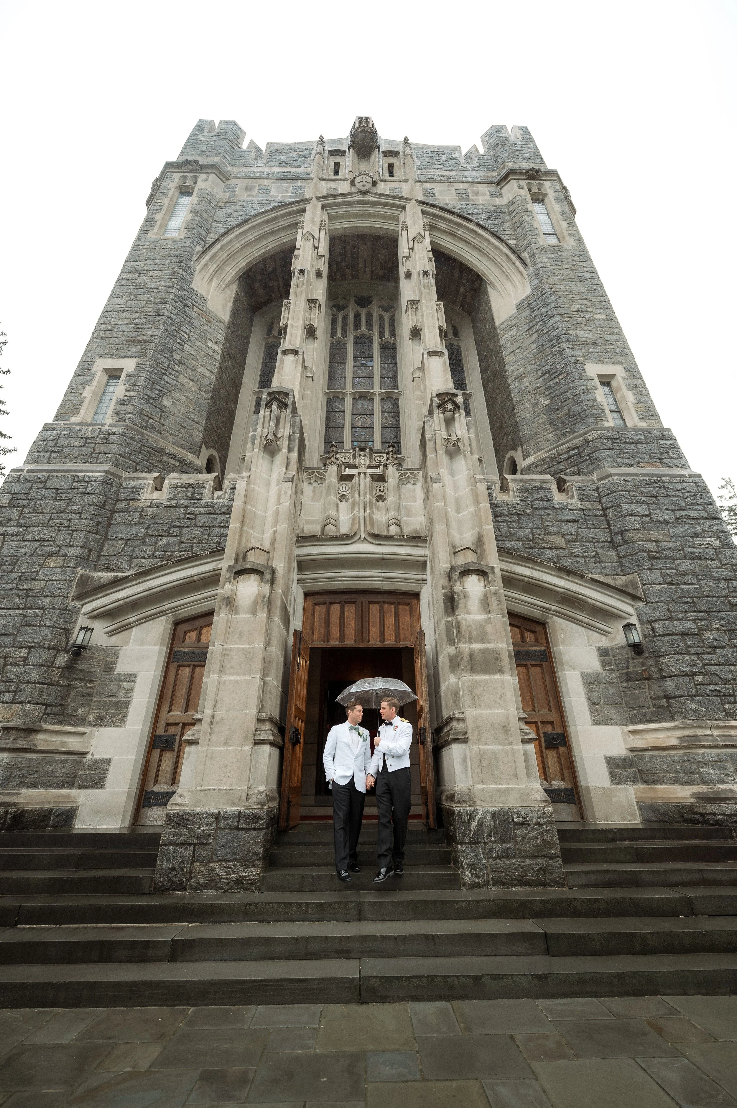 Two men in tuxedos standing under a transparent umbrella on the steps of a historic stone church, rainy weather.