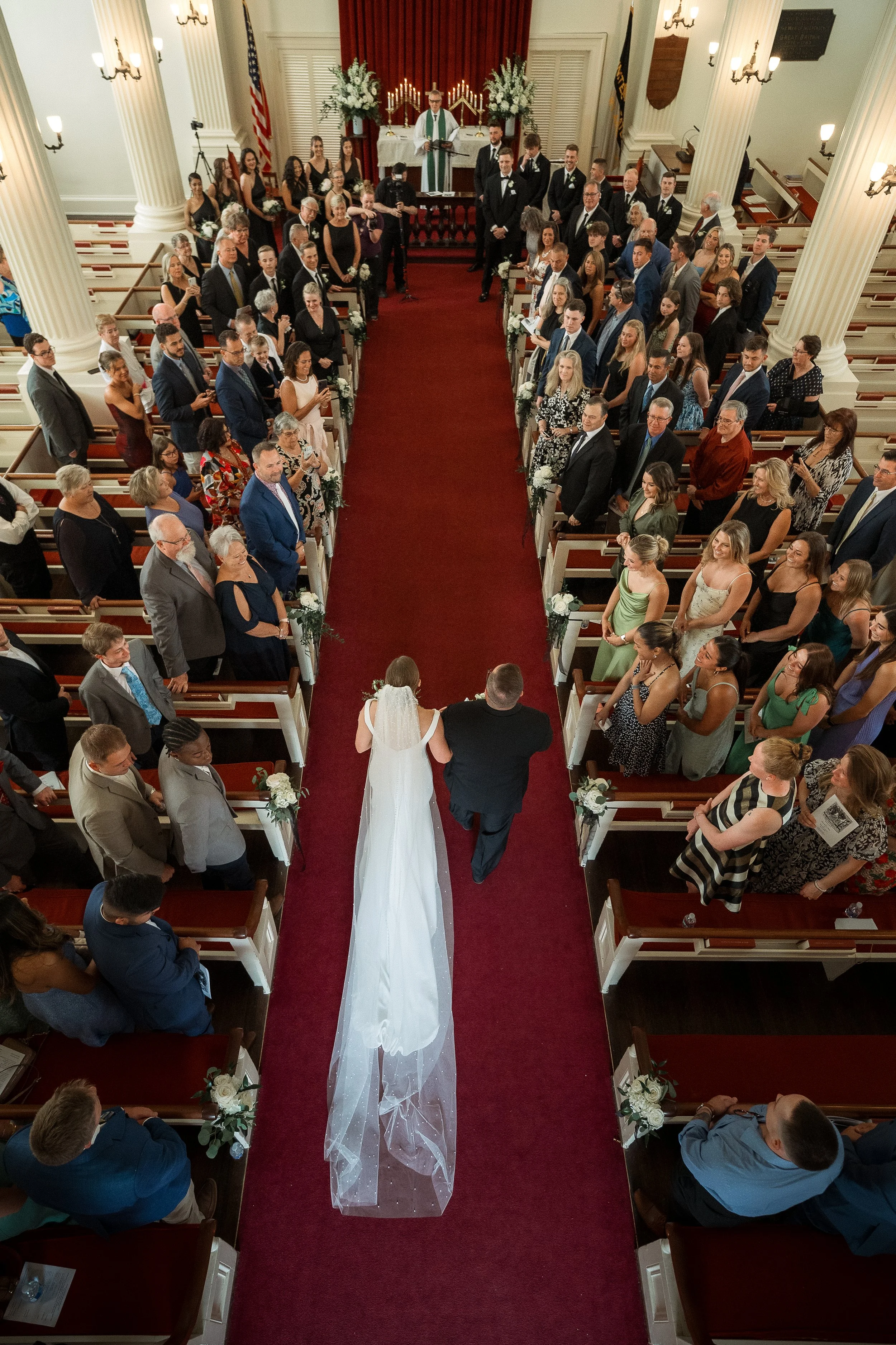 Bride walking down the aisle with her father at a church wedding, surrounded by guests seated in pews, with altar and religious decorations in the background.
