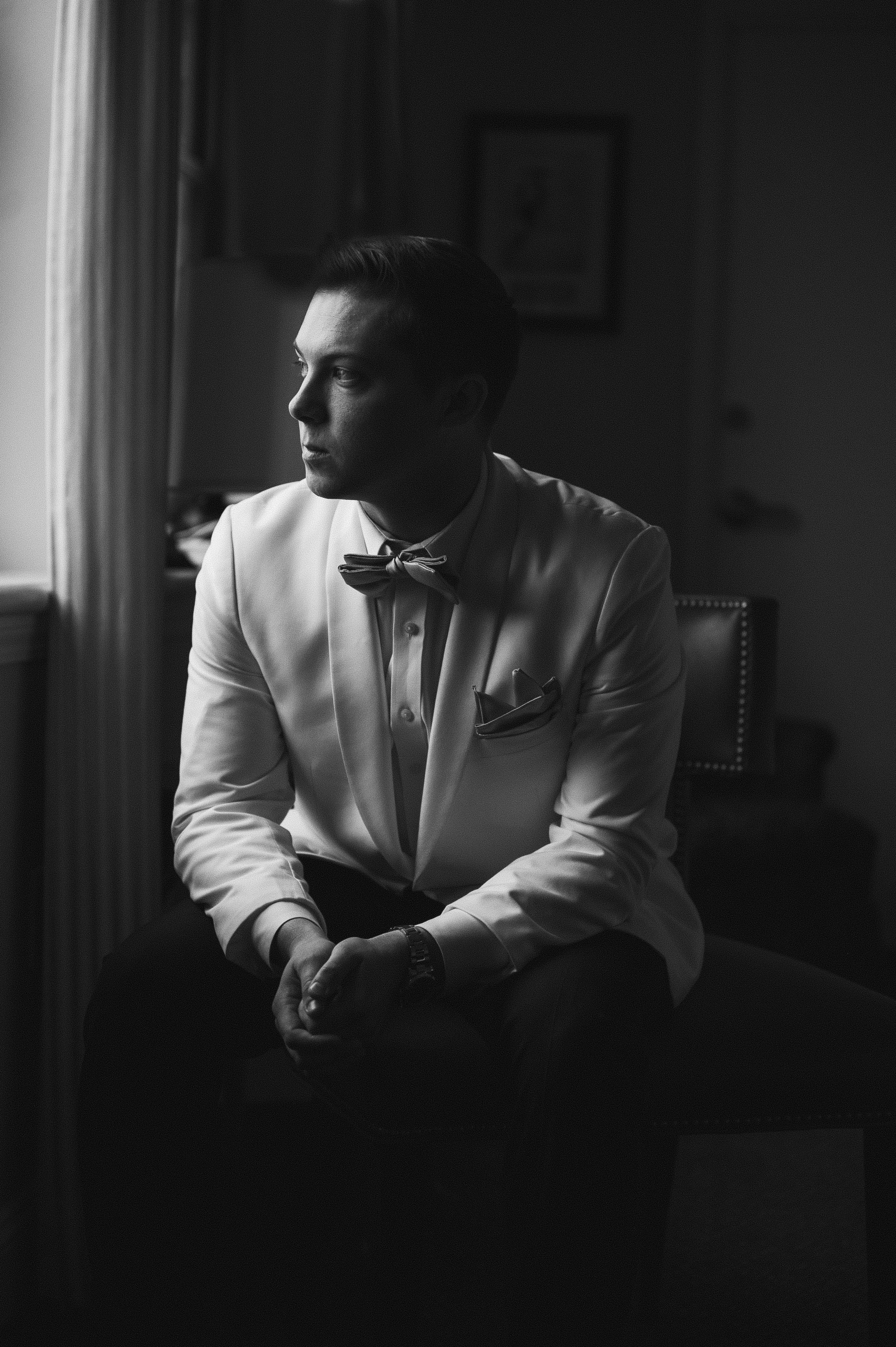 Black and white photo of a man in a tuxedo sitting on a chair near a window, looking out with natural light illuminating his face from the left.