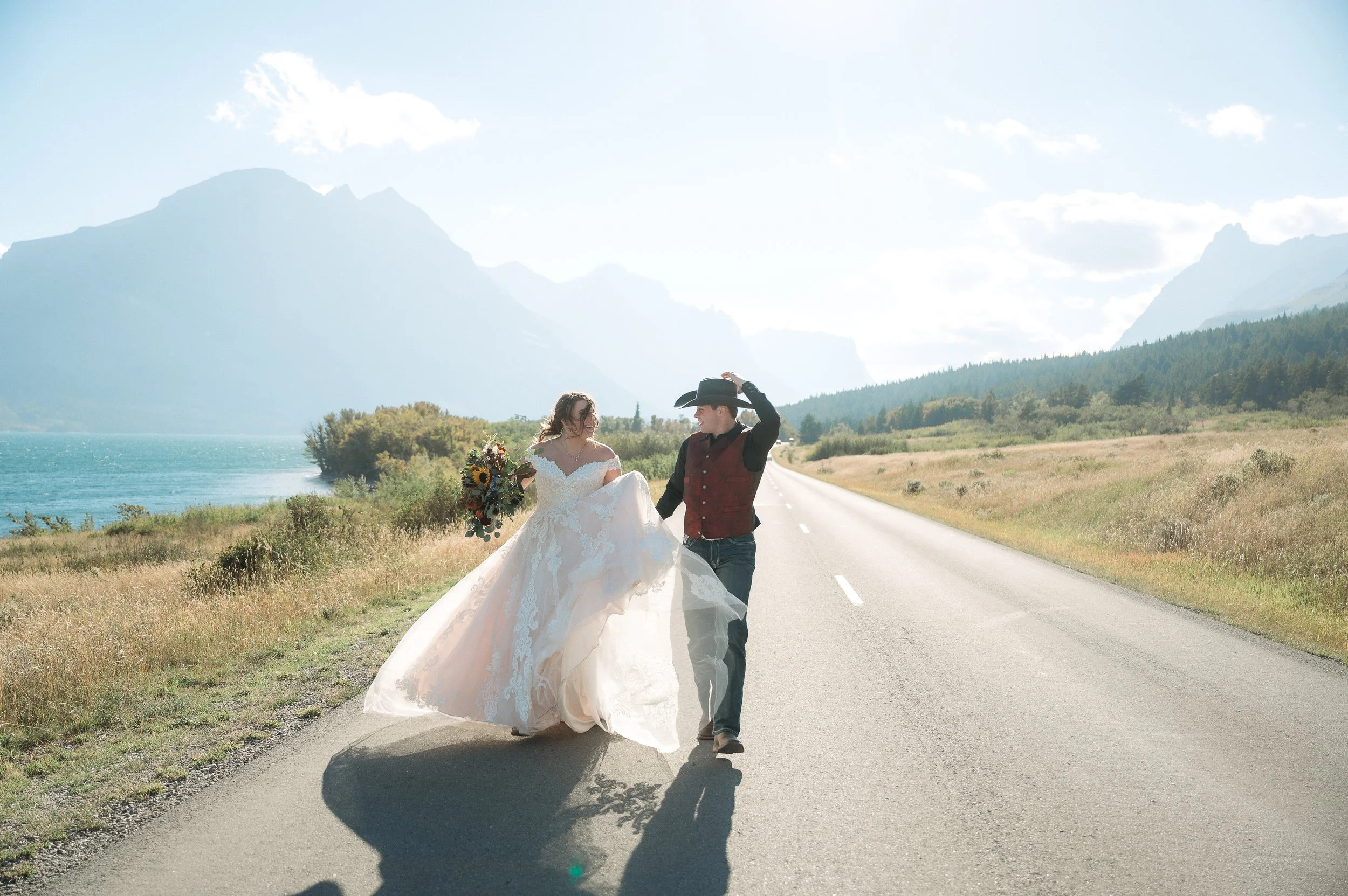 A couple in wedding attire walking on an empty road in a natural landscape with mountains and a lake in the background.