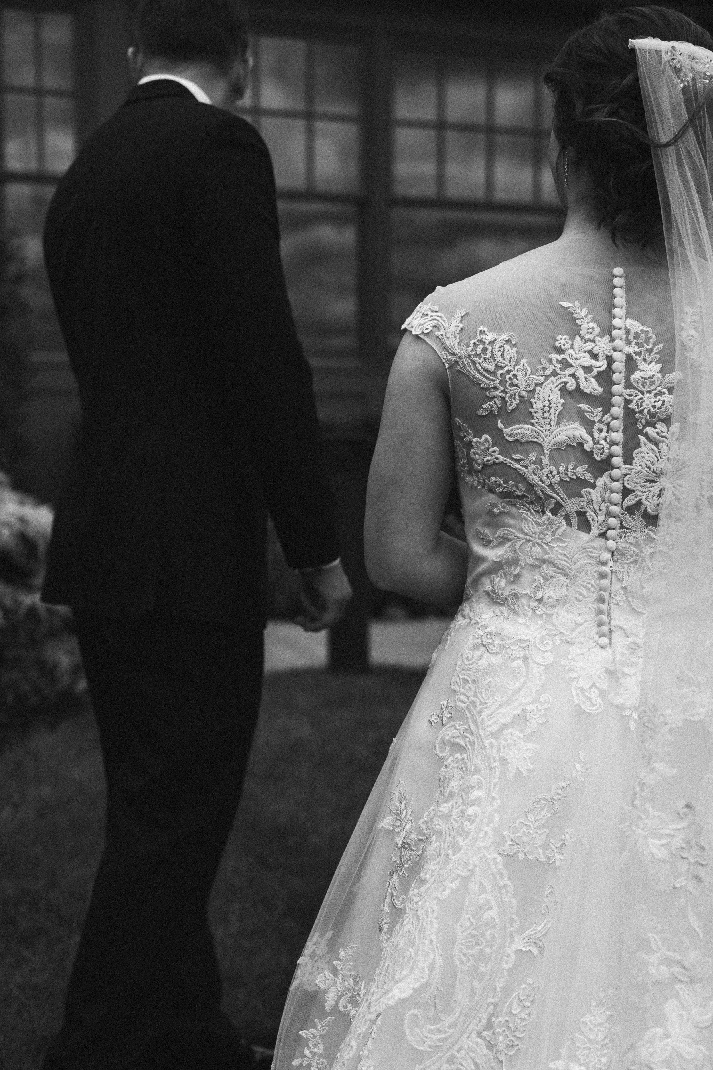 A black-and-white photo of a bride and groom holding hands during their wedding ceremony, taken from behind. The bride is wearing a lace wedding dress and a veil, and the groom is dressed in a dark suit. They are standing outdoors near a building wit