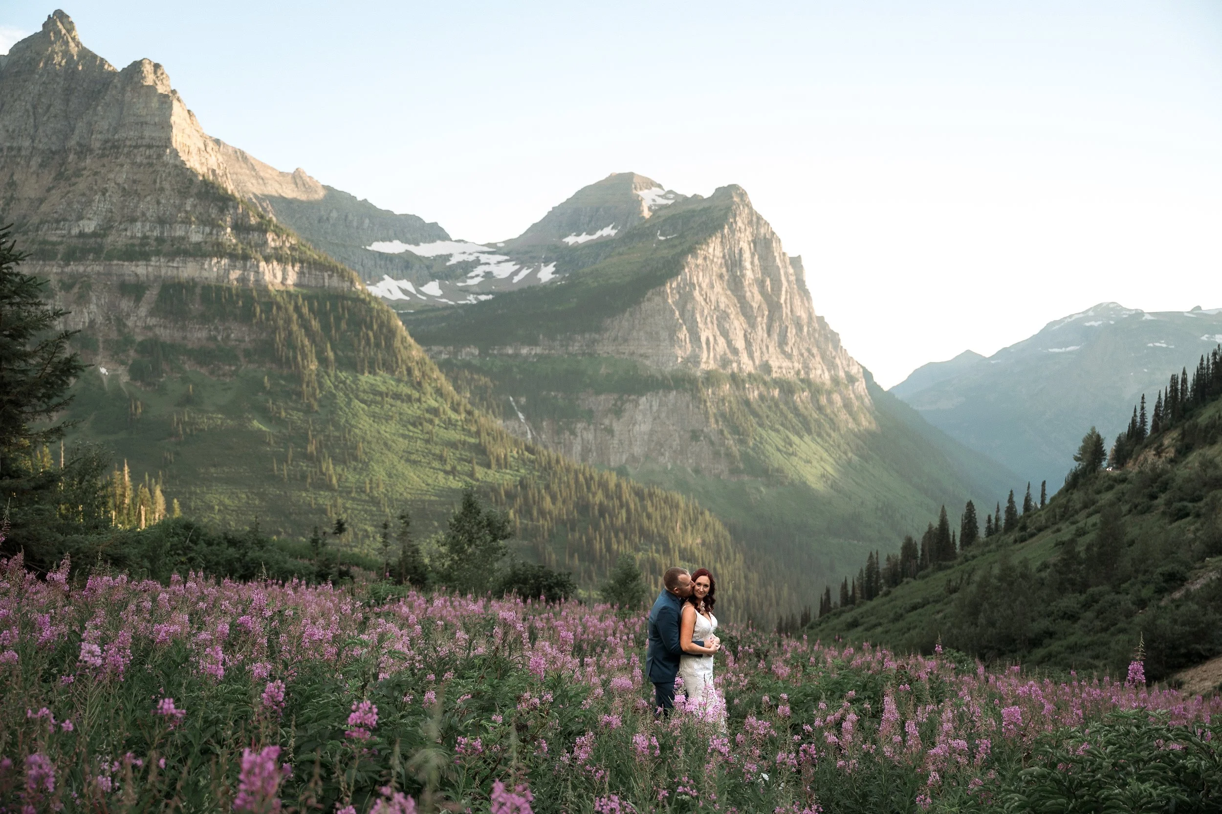 A couple in wedding attire stands in a lush field of purple wildflowers with towering green mountains in the background.
