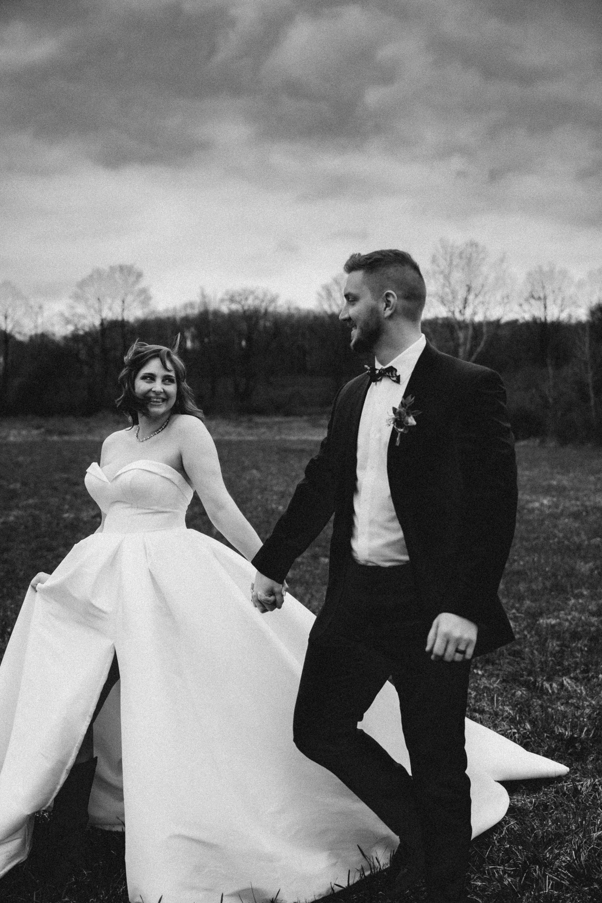 Black and white photo of a bride and groom holding hands outdoors, with trees and cloudy sky in the background.