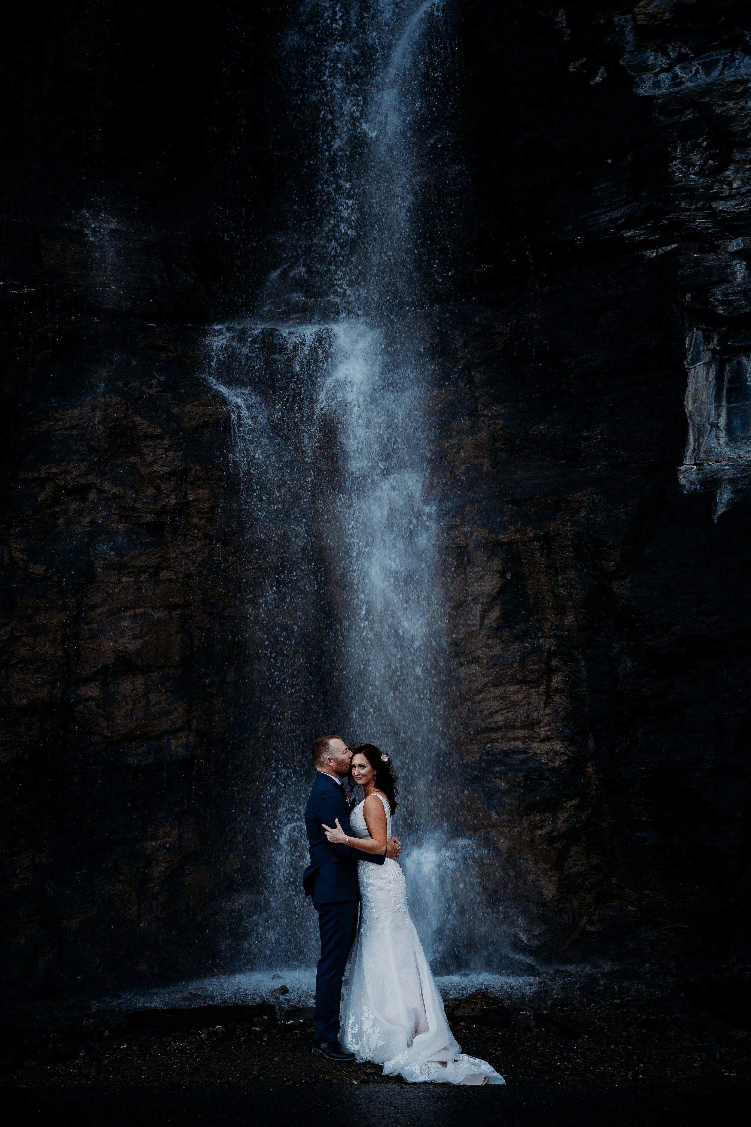 A bride and groom embrace in front of a waterfall at a dark, rocky location during their wedding.