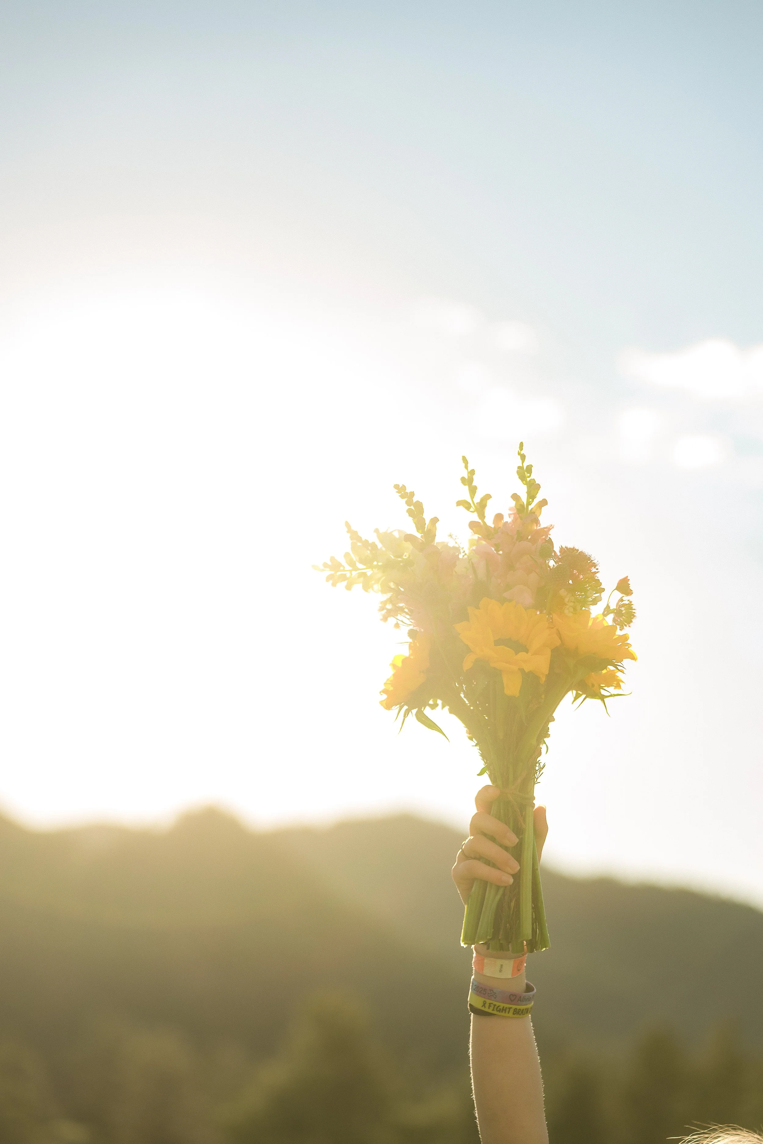 A person holding a bouquet of yellow flowers raised in the air against a bright sky with clouds and a distant landscape.