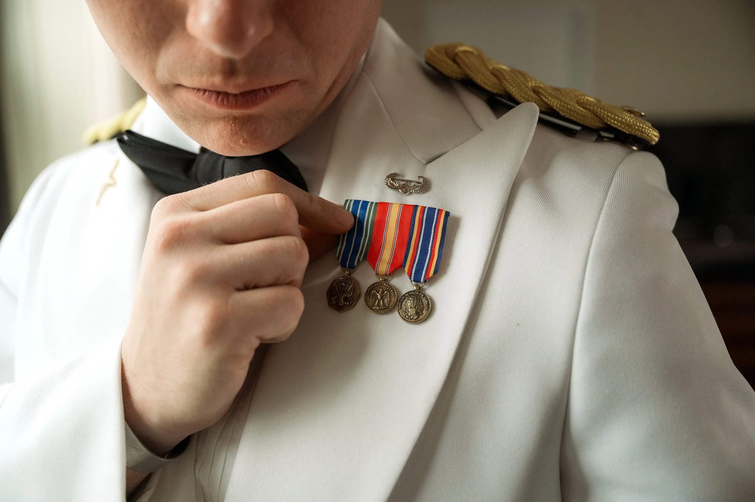 Close-up of a man in a white military uniform with gold epaulettes, adjusting or holding three medals on his chest.