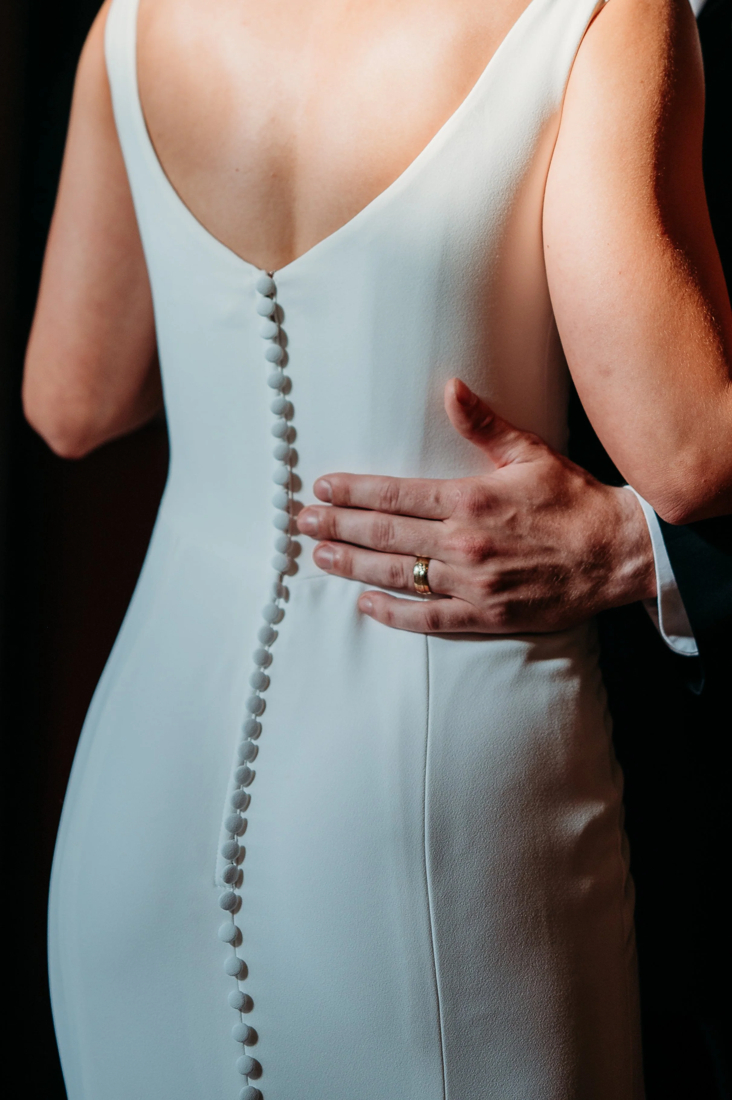 Close-up of a bride in a white wedding dress with buttons down the back, being assisted by a person wearing a dark suit, whose hand is resting on the bride's waist with a wedding ring visible.