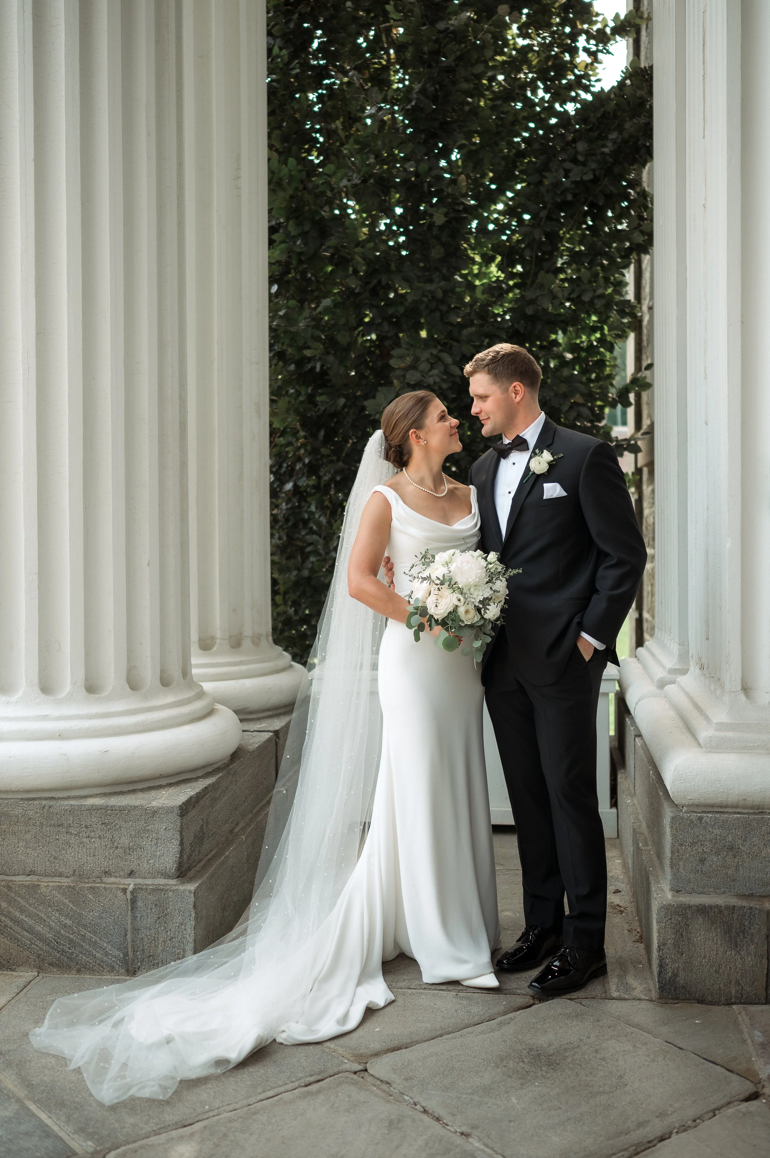 A bride and groom standing close together under a columned structure on their wedding day, gazing at each other, with the bride holding a bouquet of white flowers and wearing a white gown and veil, and the groom in a black tuxedo.